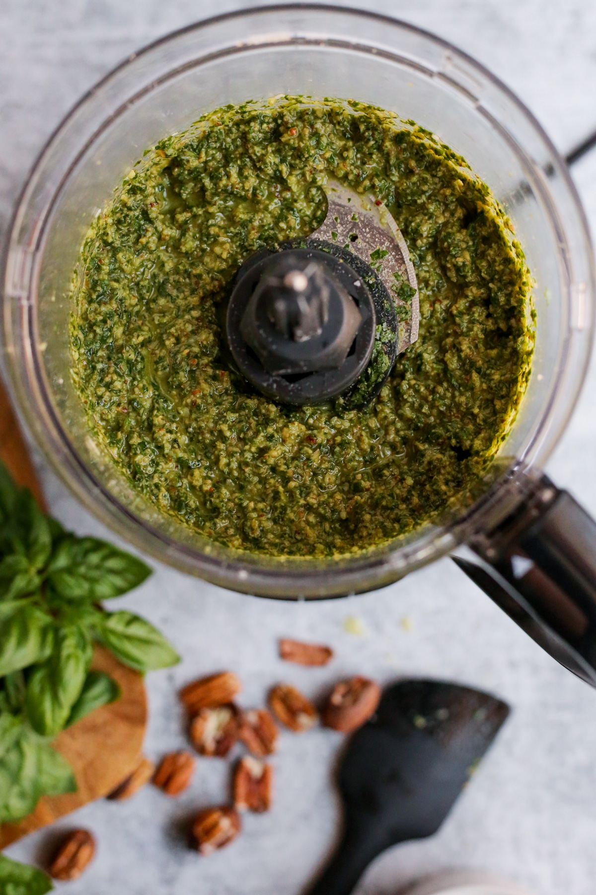 Overhead view of a fully blended batch of pecan pesto without pine nuts in the bowl of a food processor, set up on a kitchen countertop with the lid removed to show the chunky consistency after the olive oil is added