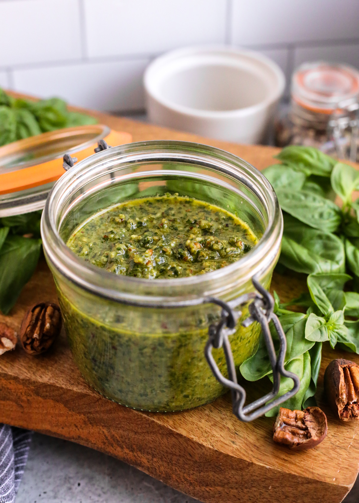 A view of a freshly made batch of homemade basil pecan pesto in a glass jar, displayed on a wooden serving tray on a kitchen countertop and surrounded by shelled pecans and fresh basil leaves