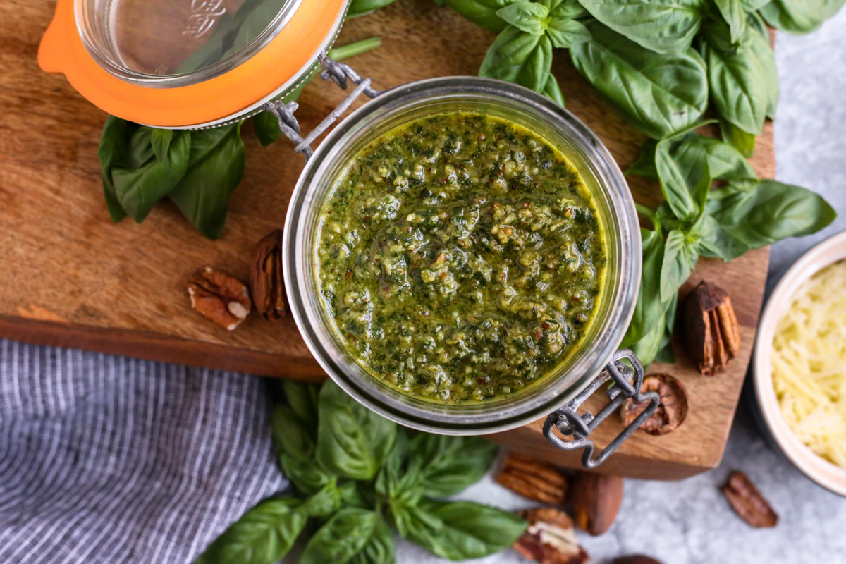 Landscape image with an overhead view of homemade basil pecan pesto without pine nuts, stored in a glass jar with a hinged lid, open to display the vibrant green pesto sauce while surrounded by additional ingredients