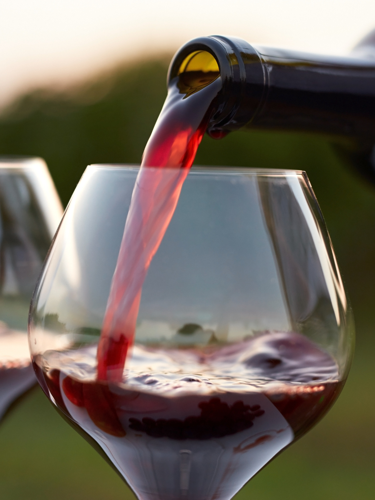 A close cropped image of a large wine glass with a bottle of red wine being poured into it in an outdoor setting. A second wine glass is partially visible in the background