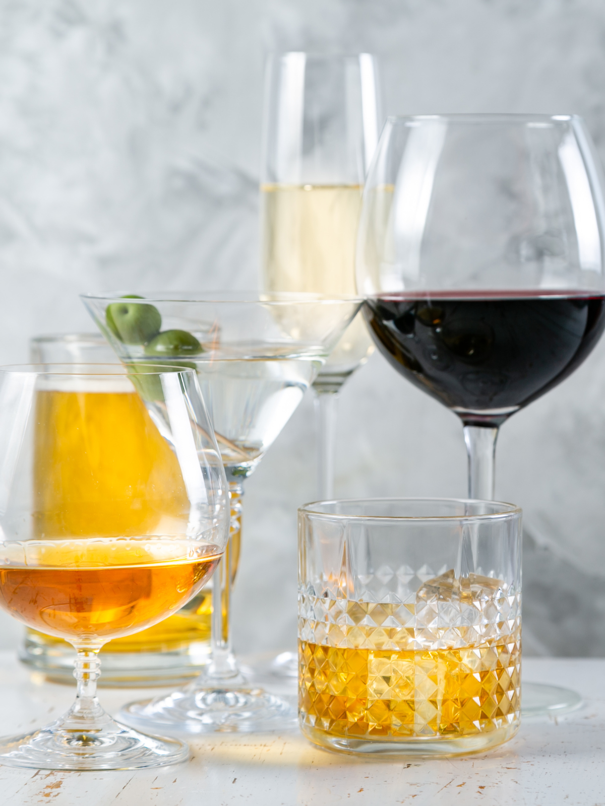 A variety of liquor, beer, wine, and spirits are arranged in glasses on a grey kitchen countertop, showcasing the variety in drink sizes that equal a standard drink