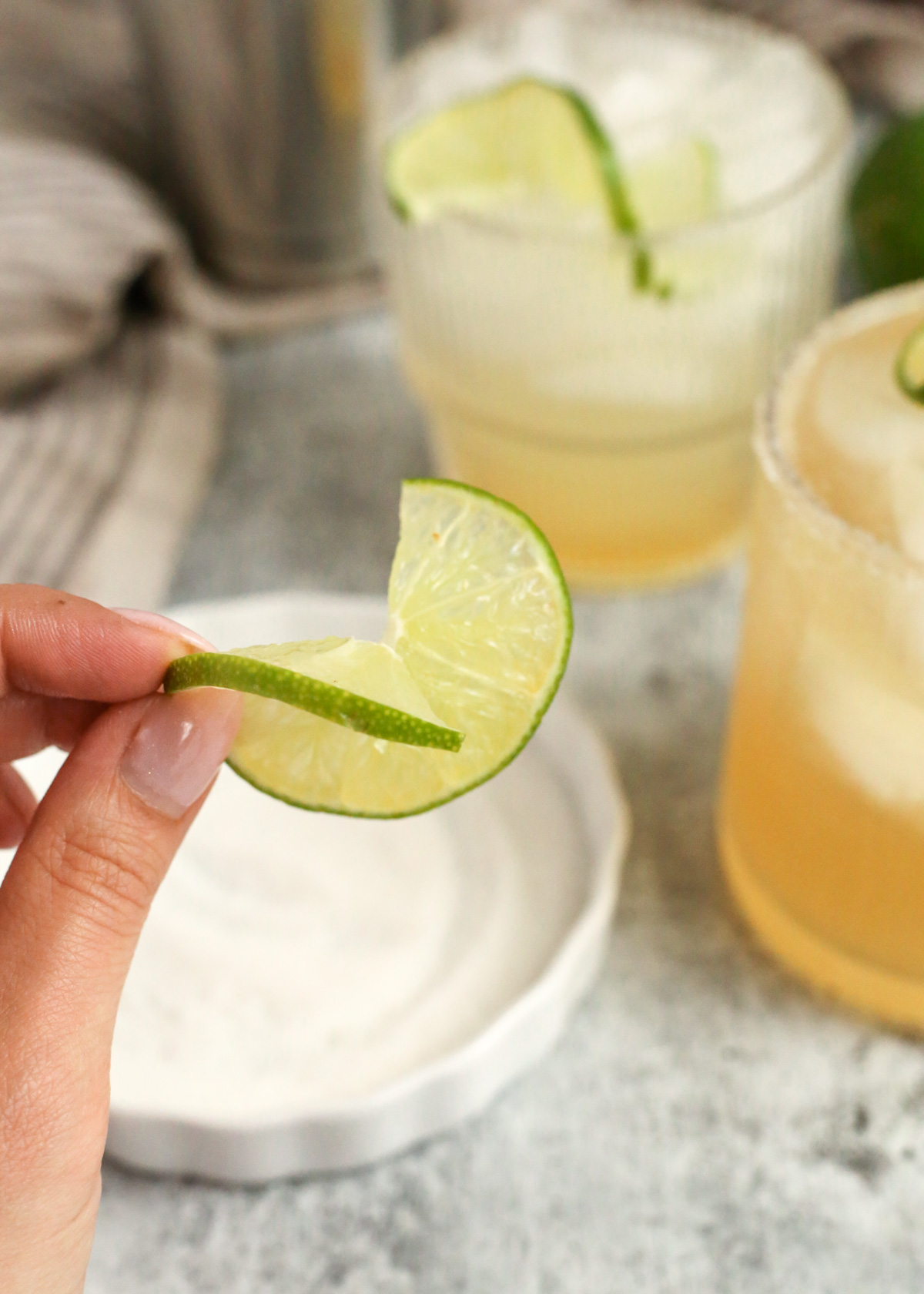 A woman's hand holds a thin slice of lime that's been cut halfway through and twisted to form a spiral shape to garnish a yuzu margarita