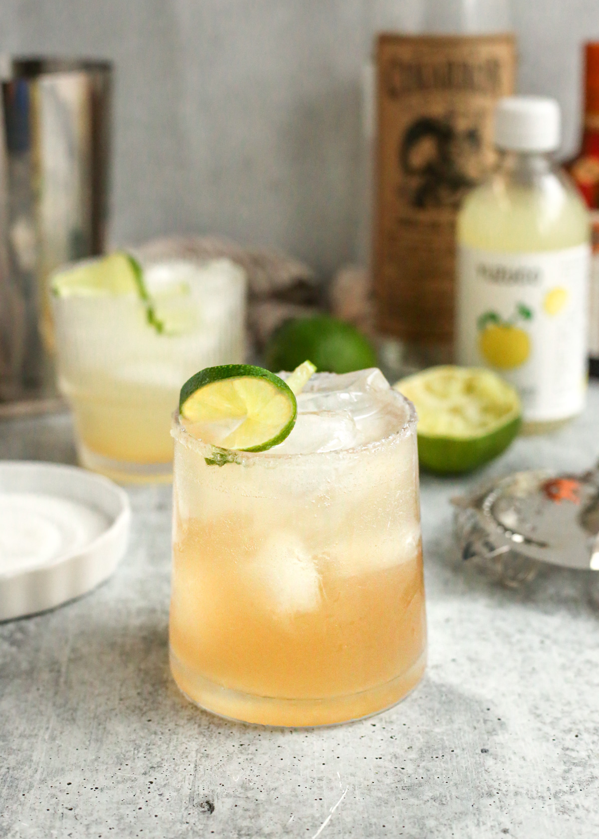 A yuzu margarita served in a clear glass on a kitchen countertop with a silver stainless cocktail shaker and various liquor bottles in the background. The drink is garnished with a lime wheel and the glass is frosted to make the drink look extra refreshing