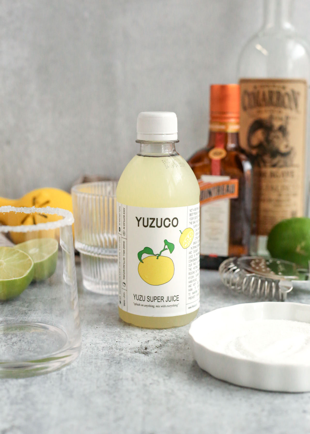 A bottle of light yellow-green yuzu juice sits on a kitchen counter surrounded by the other ingredients for a yuzu margarita. The label reads Yuzuco yuzu super juice