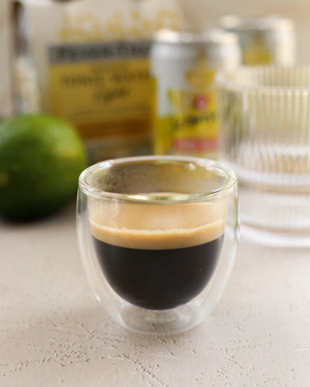 A double shot of freshly brewed espresso in a clear, double-walled glass sits in front of cans of tonic water 