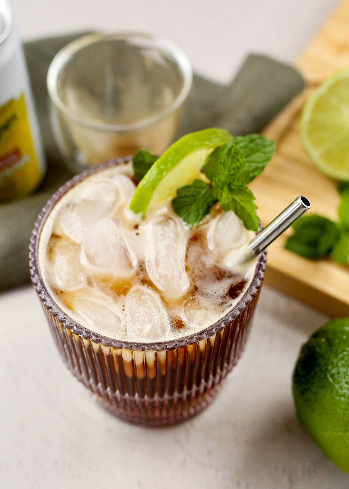 Angled view into a short rippled glass that's filled with ice cubes and and iced espresso tonic, garnished with a lime wedge and mint leaf with a short silver metal straw resting against the right side of the glass