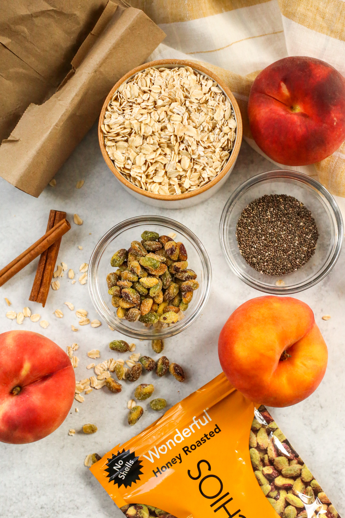 Overhead view of various sizes of ramekins and small glass prep bowls containing old-fashioned oats, chia seeds, and pistachios, with cinnamon sticks and fresh peaches included nearby