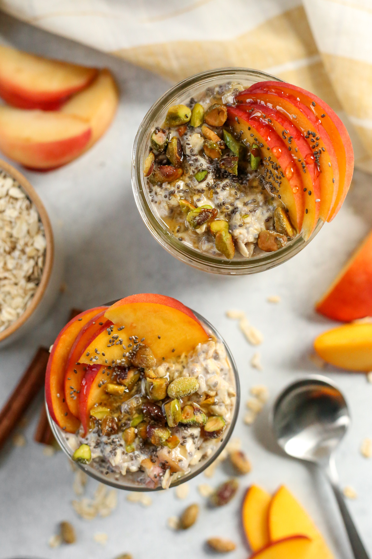 Overhead view of two jars of Peaches and Cream Overnight Oats, beautifully garnished with sliced peaches, pistachios, chia seeds, and a drizzle of honey, with extra oats and peach slices scattered nearby