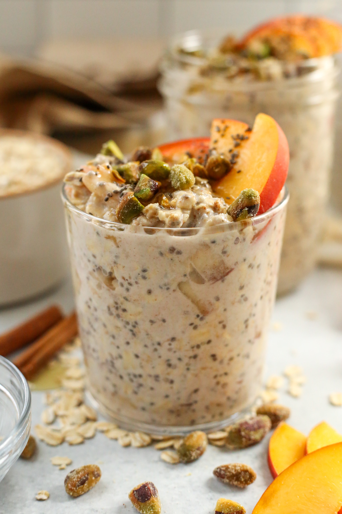 A glass jar full of creamy, thick overnight oats is topped with fresh peach slices, shelled pistachios, and chia seeds, served on a kitchen countertop with extra ingredients scattered around it 
