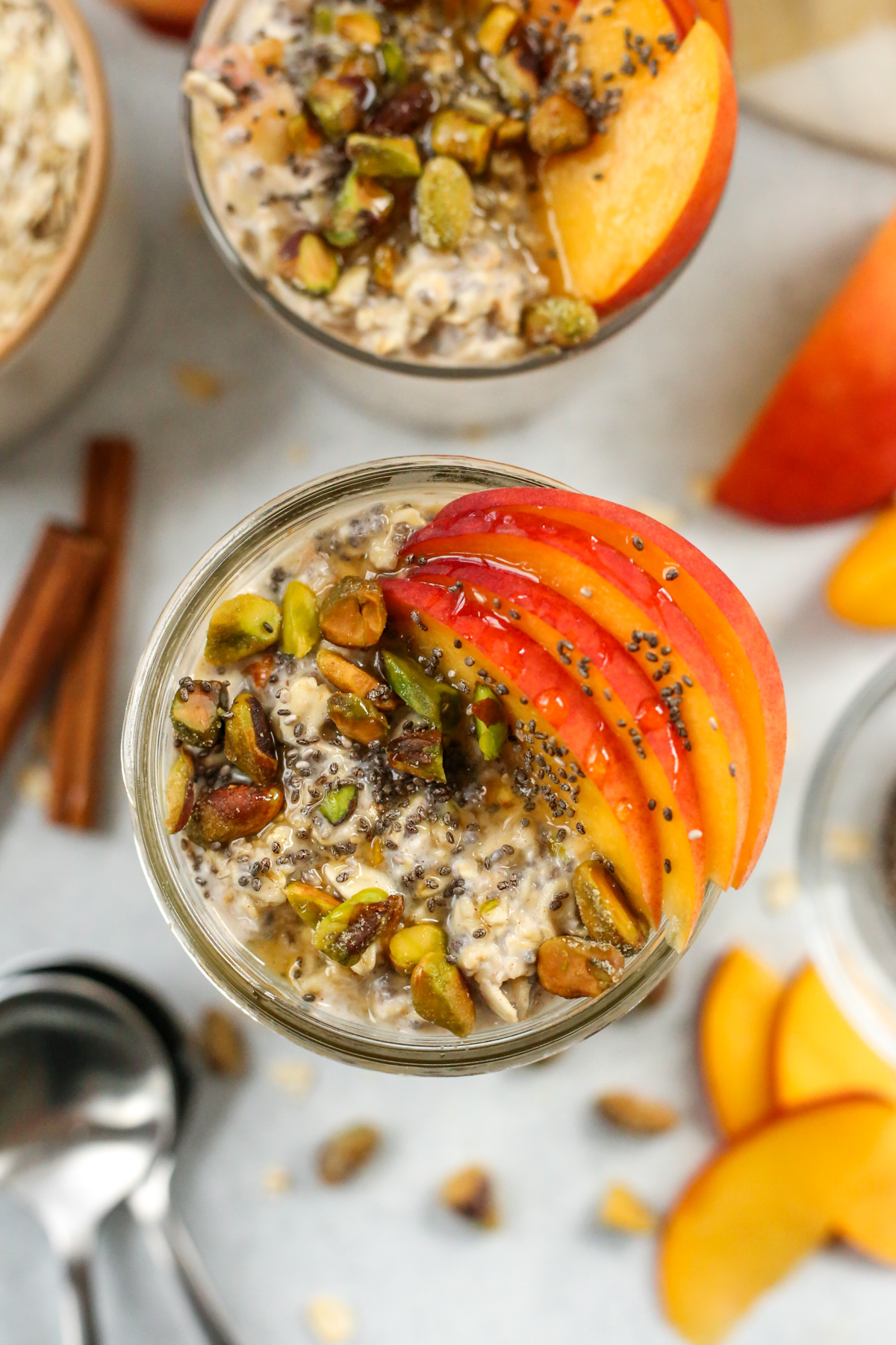 Close up view of thin slices of peaches resting on top of a mason jar containing overnight oats, with a few drops of honey freshly drizzled on top of them, along with some shelled pistachios and chia seeds for garnish
