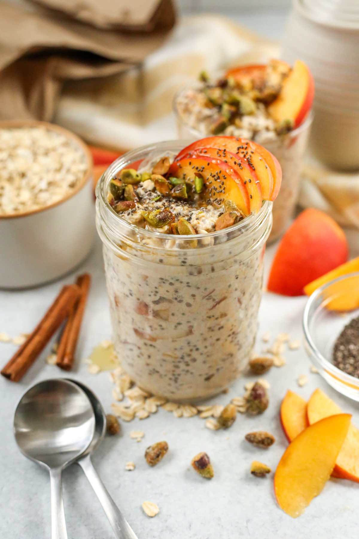 A styled shot of a serving of peaches and cream overnight oats served in a mason jar in a kitchen scene, with serving spoons, cinnamon sticks, extra ingredients, and fresh peaches displayed around it