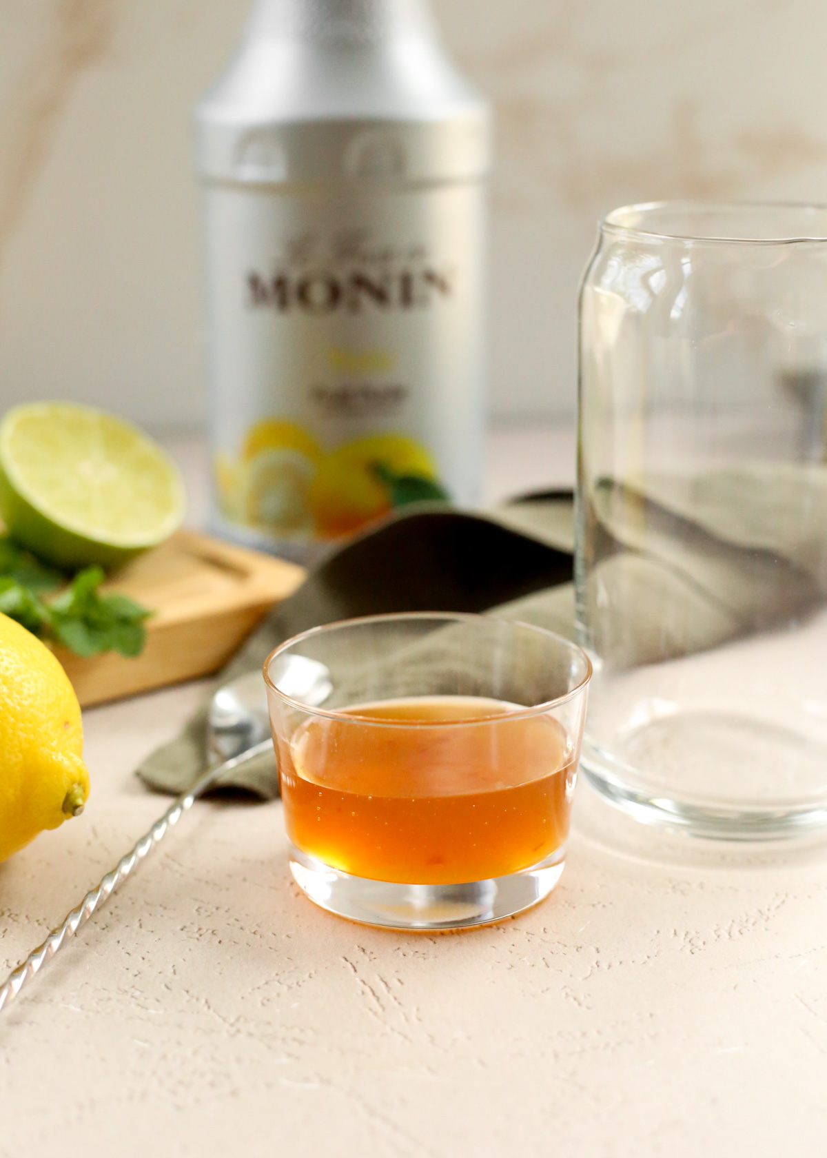 A small glass ramekin is filled halfway with vibrant orange-colored yuzu puree. A bar spoon, empty glass, and citrus fruits are scattered nearby