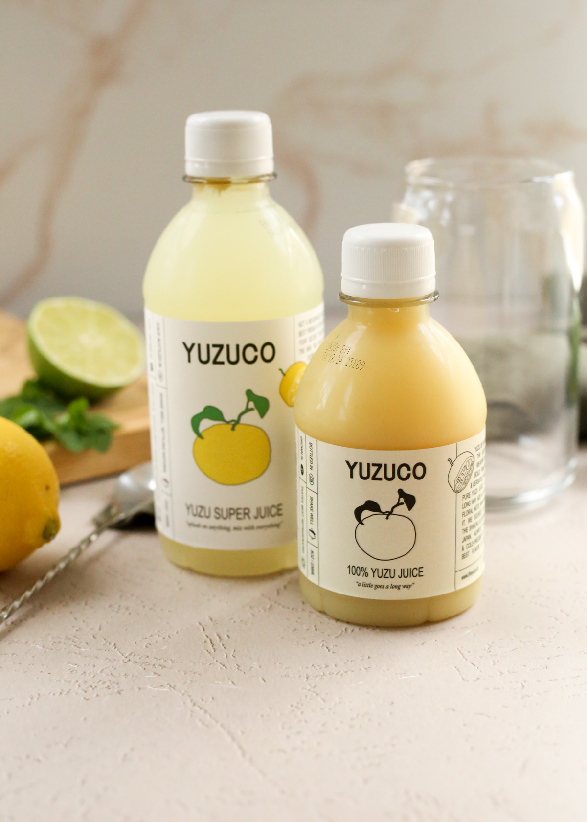 Two bottles with Yuzu Co labels sit on a kitchen countertop, with a smaller bottle of 100% yuzu juice displayed slightly in front of a larger bottle of yuzu super juice. Both bottles are filled with a light yellow citrus juice with white caps fastened