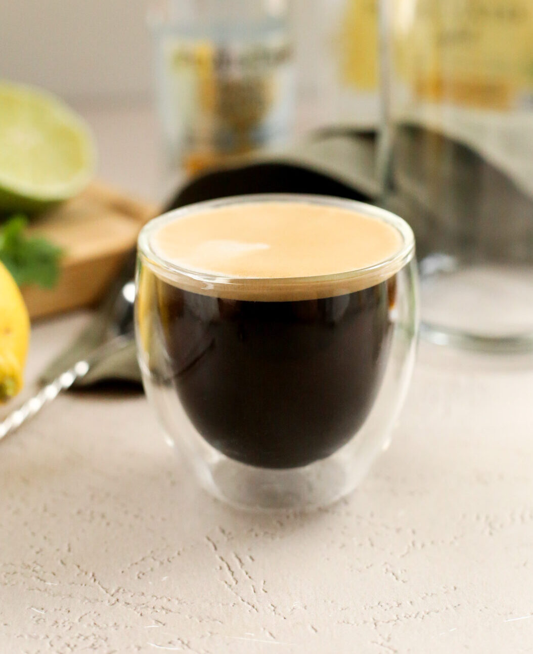 A shot of espresso in a double walled clear glass, served on a kitchen countertop in warm afternoon light with a silver metal bar spoon and empty glass in the background