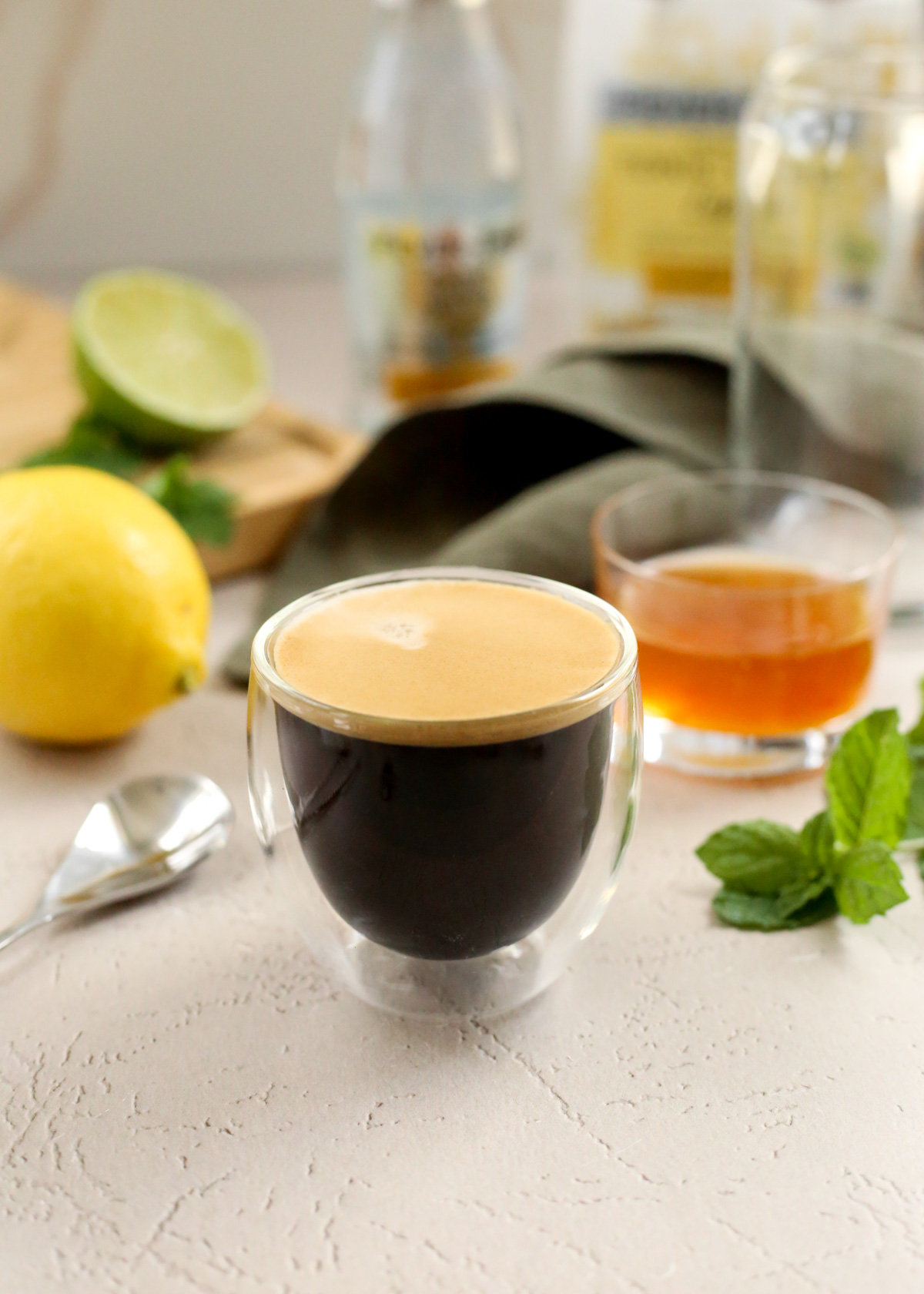 A double shot of espresso in a double walled espresso glass sits on a kitchen countertop with citrus fruits, a small ramekin of yuzu puree, and various bottles of tonic water in the background