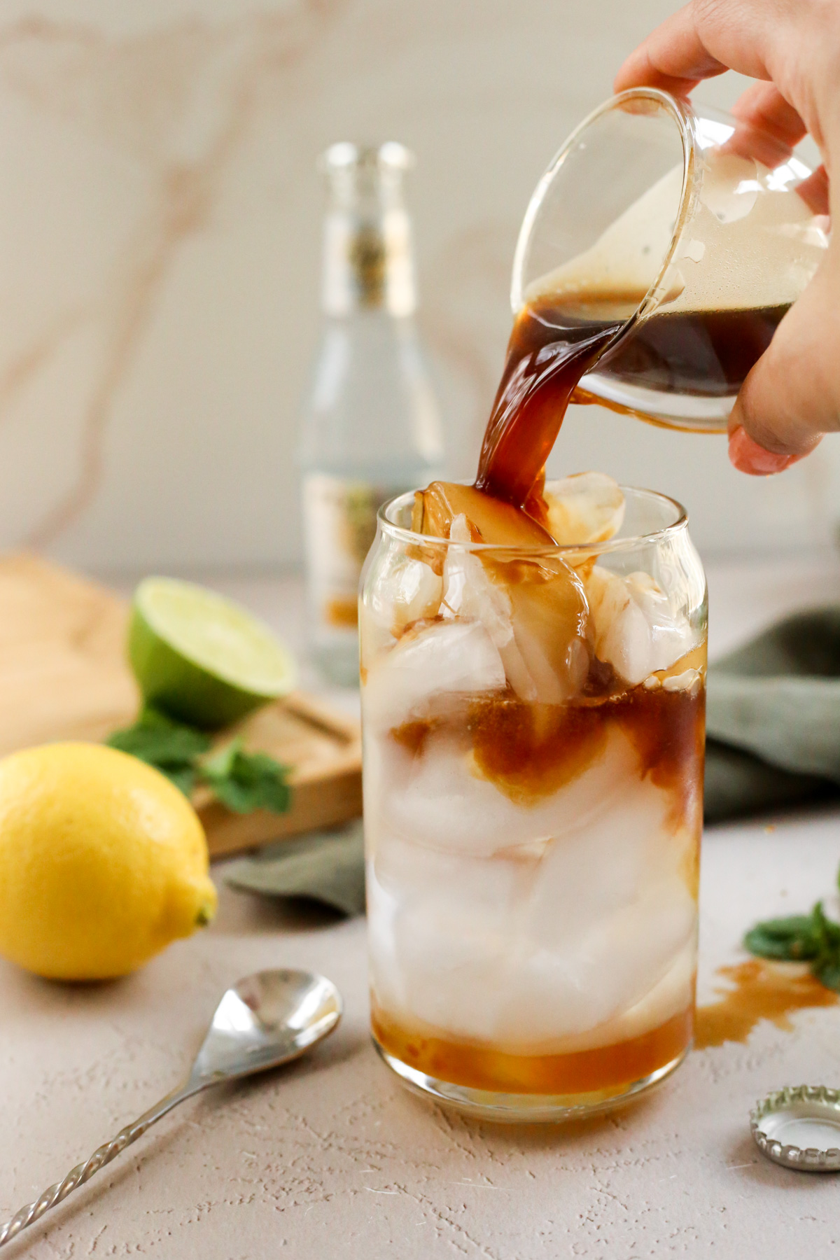 A woman's hand pours a shot of espresso over ice in a clear glass, with the espresso mixing with the tonic water and yuzu puree already in the glass