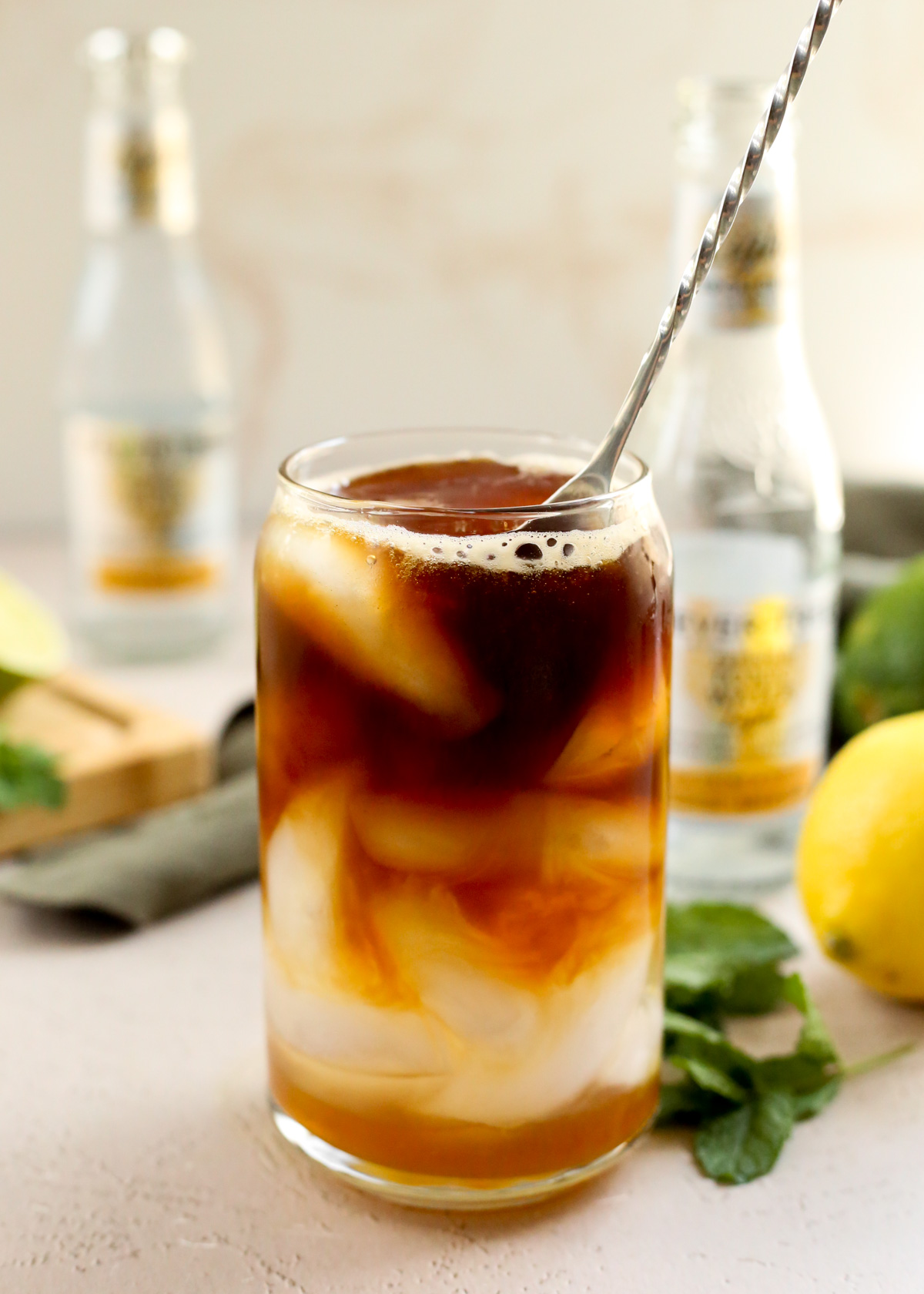 A refreshing summer coffee drink in a clear glass, with layers of yuzu puree on the bottom, tonic water, and espresso with ice cubes, and a cocktail stirrer resting in the glass as if about to break up the layers with mixing