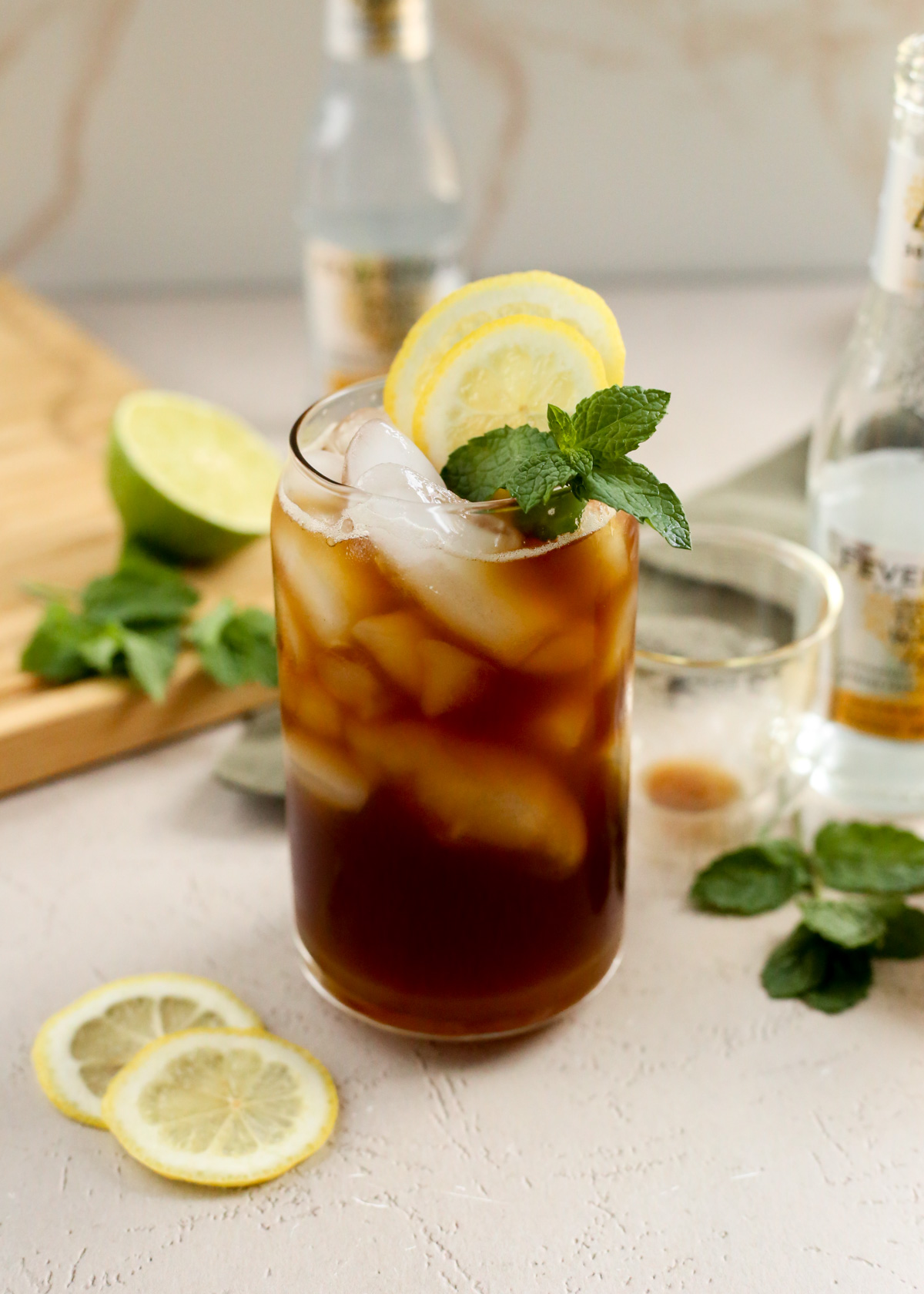 A yuzu espresso tonic is served in a clear glass with many ice cubes and lemons wheels plus a sprig of fresh mint as a garnish, which makes it look like a refreshing summer coffee drink for a hot day