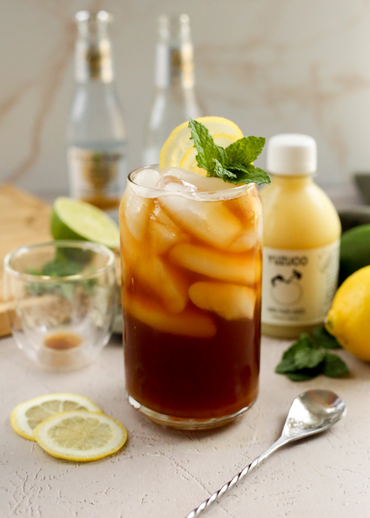 A yuzu espresso tonic is served in a clear glass with ice cubes, lemon wheels, and fresh mint as garnish, with an empty espresso glass and bottle of yuzu juice visible in the background. It looks like a refreshing summer coffee drink for a hot day