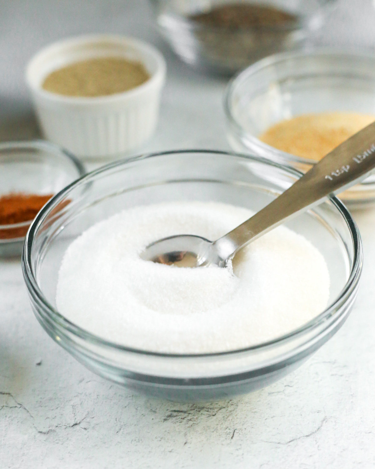 A silver measuring spoon rests in a pile of MSG, which looks like a pile of salt, contained in a small glass prep bowl and surrounded by other dried spices and seasonings on a kitchen countertop