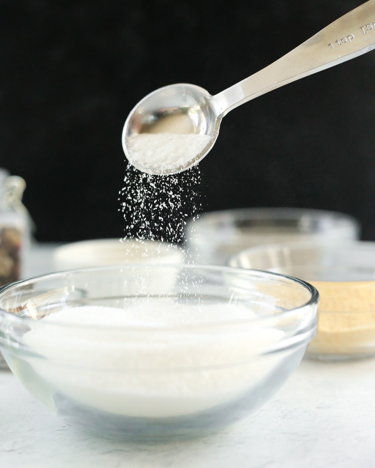 A silver measuring spoon pours MSG into a small glass prep bowl, in front of a dark background showing the individual white crystals as they fall