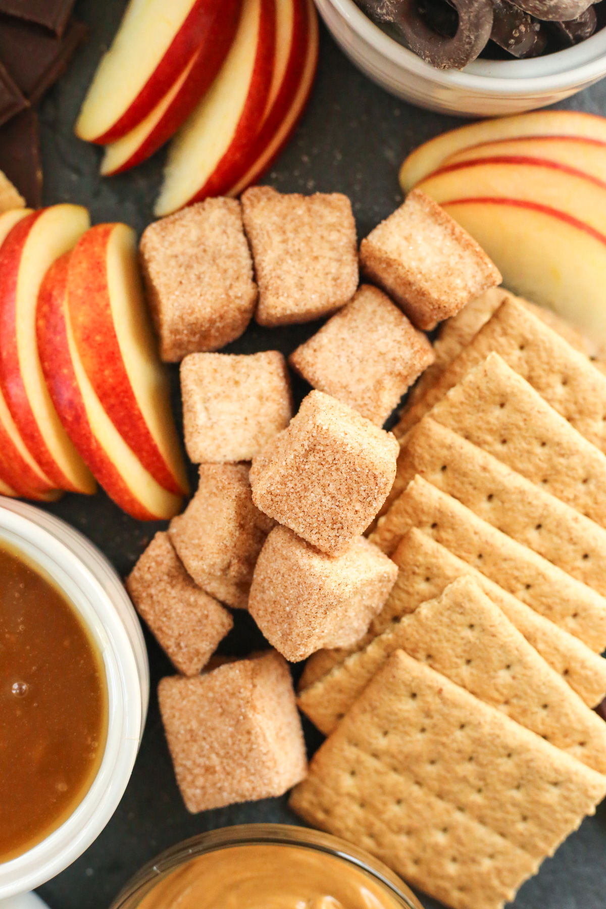 Close up overhead shot of small square marshmallows, dusted and coated with cinnamon sugar, alongside graham cracker squares, sliced apples, and a small ramekin of caramel sauce