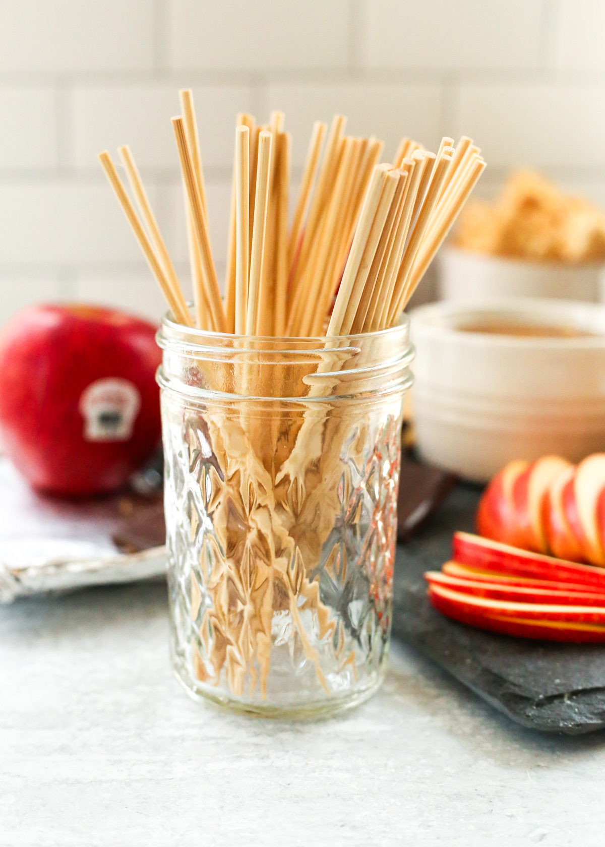 A small clear mason jar holds mini kebab skewers, intended to be used to toast a marshmallow for a s'more, with a dessert board that includes apple slices visible in the background