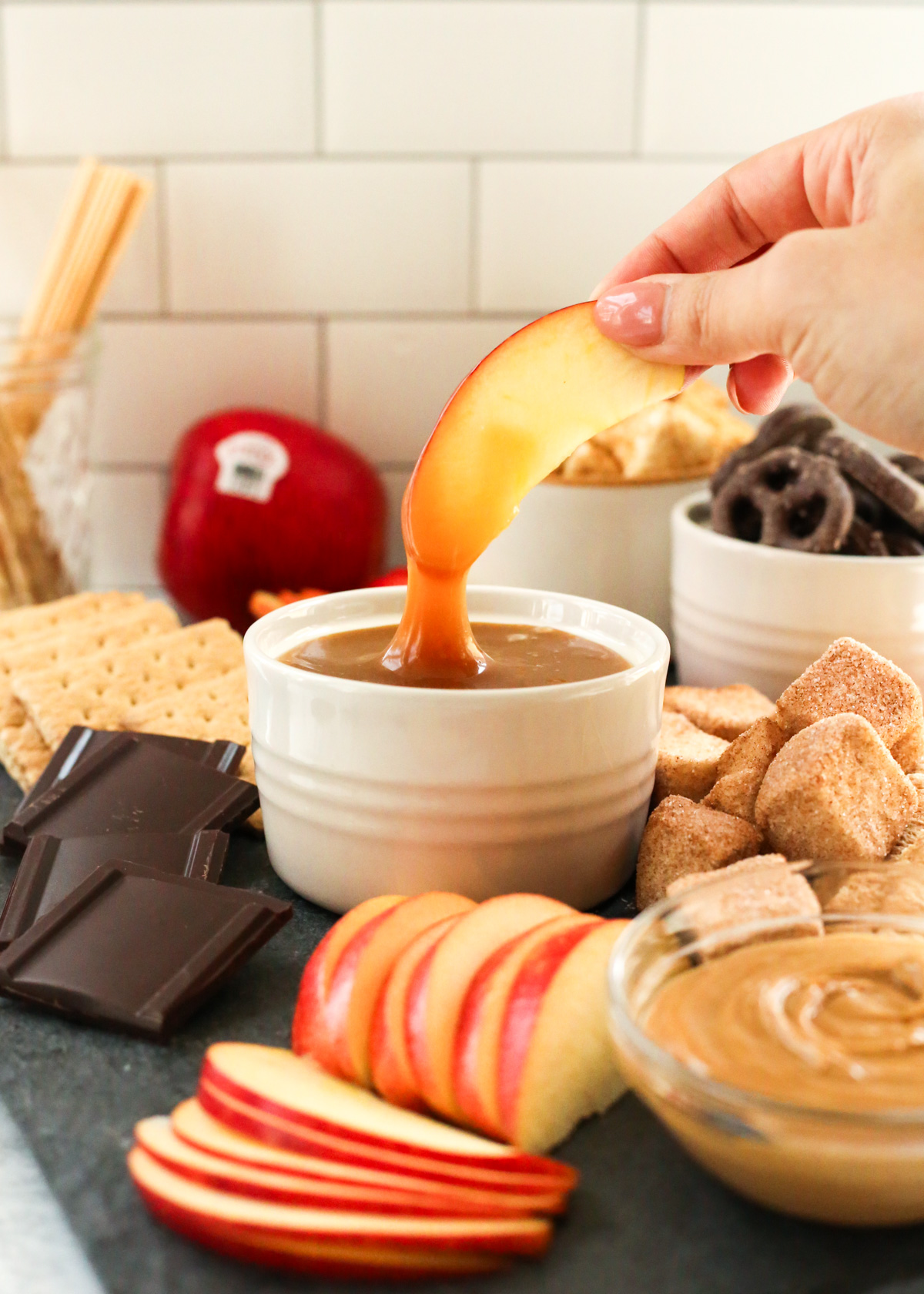 A woman's hand dips an apple slice into a small ramekin full of caramel sauce, with more apple slices and s'mores ingredients scattered on a dessert board in the background