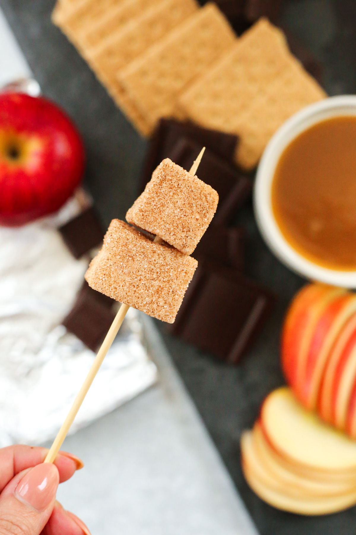 A woman's hand holds a mini wooden skewer pierced through two of the square marshmallows coated with cinnamon sugar, with a caramel apple dessert board in the background