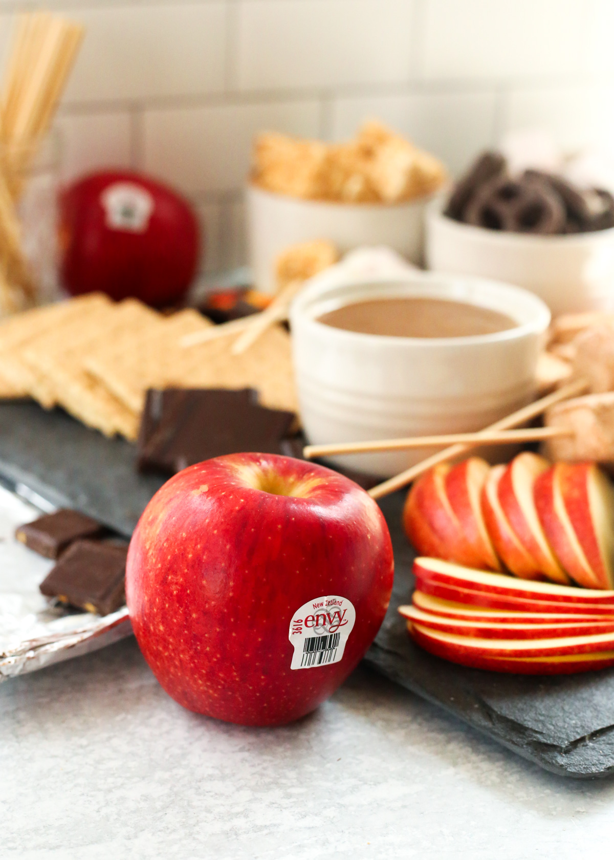 A centered shot of an Envy apple, with bright red and yellow skin, sitting in front of a dessert board with chocolate, chocolate covered pretzels, marshmallows, graham crackers, and various other ingredients in the background