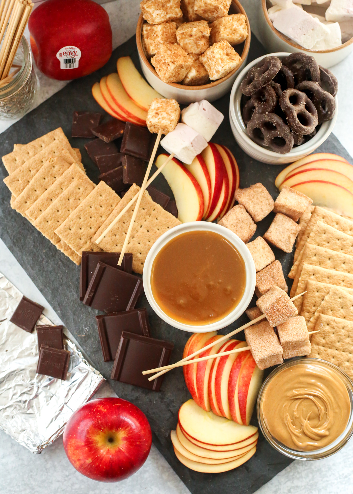 Overhead view of a caramel apple s'mores dessert board, served on a piece od dark grey slate, with various ingredients for caramel apples and s'mores arranged randomly 