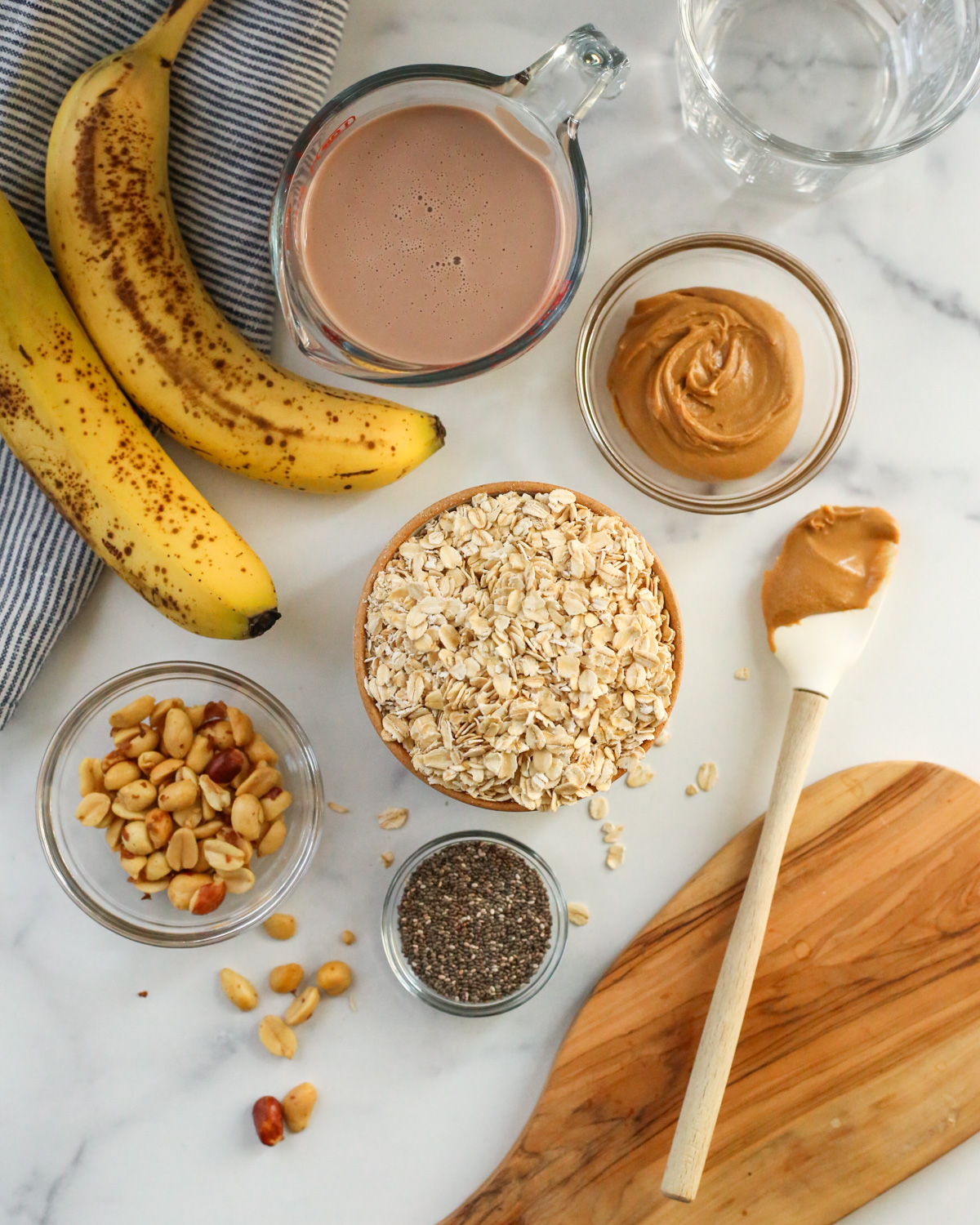 Overhead view of an arrangement of ingredients for chocolate banana overnight oats, including a pair of ripe, unpeeled banana, a measuring cup full of chocolate milk, and small ramekins holding oats, peanuts, peanut butter, and chia seeds