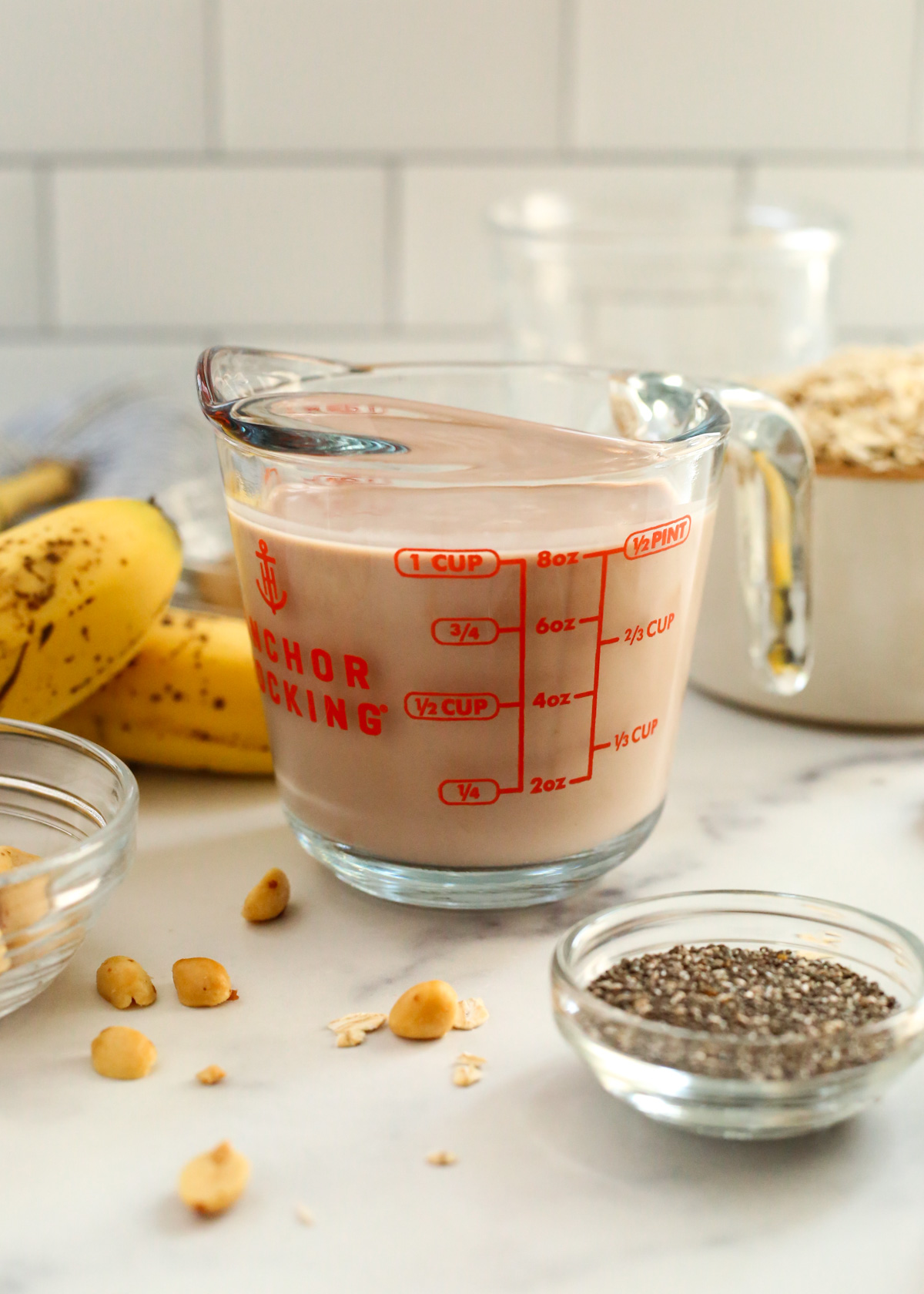 A glass measuring cup is filled with chocolate milk, angled to allow the red lettering and lines to be seen. Random ingredients for an overnight oats recipe are scattered nearby 