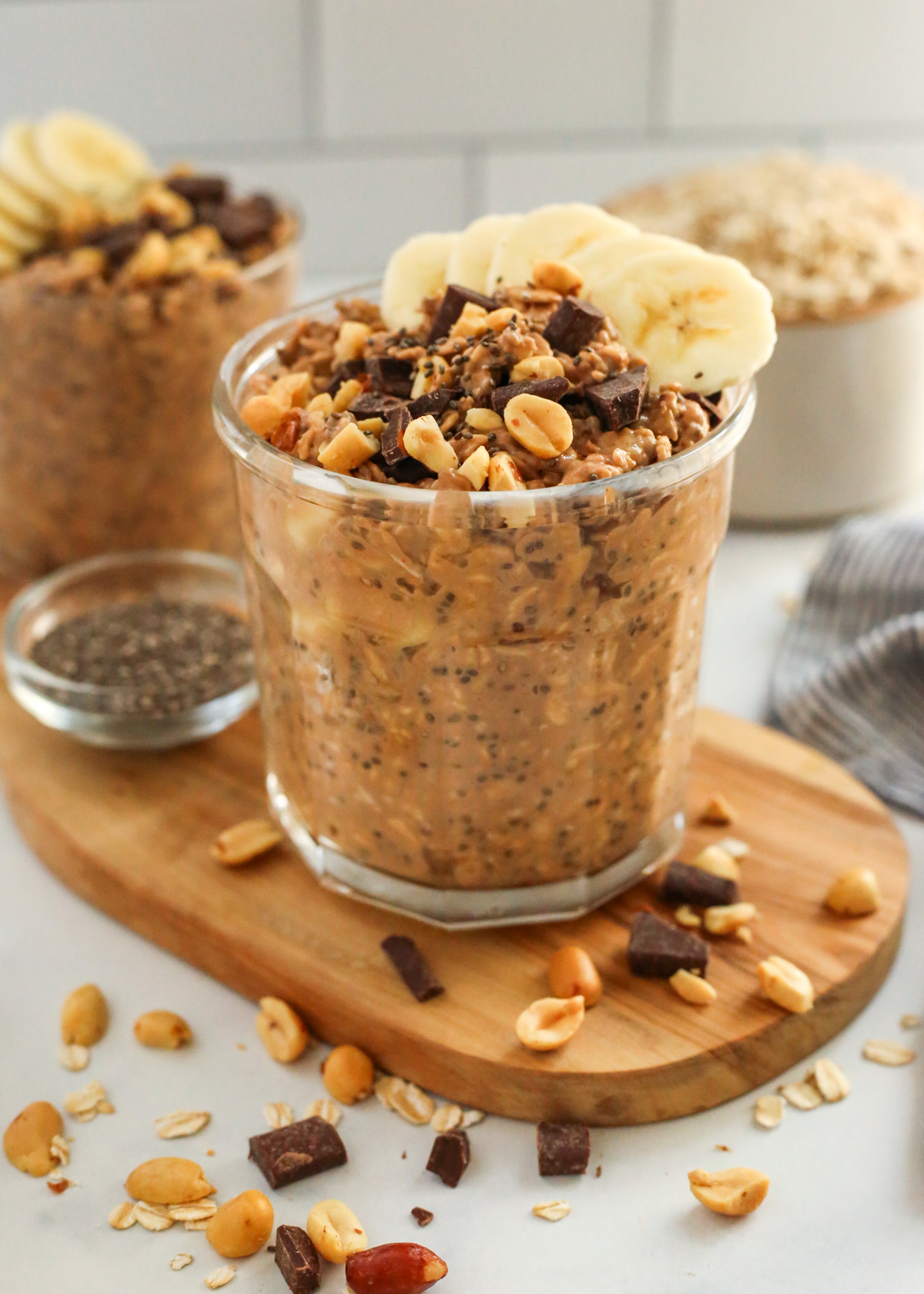 Two jars of chocolate banana overnight oats are displayed in a kitchen scene, with one jar positioned closer to the view on a wooden serving board. Both are topped with pieces of peanuts, chocolate chunks, and sliced bananas