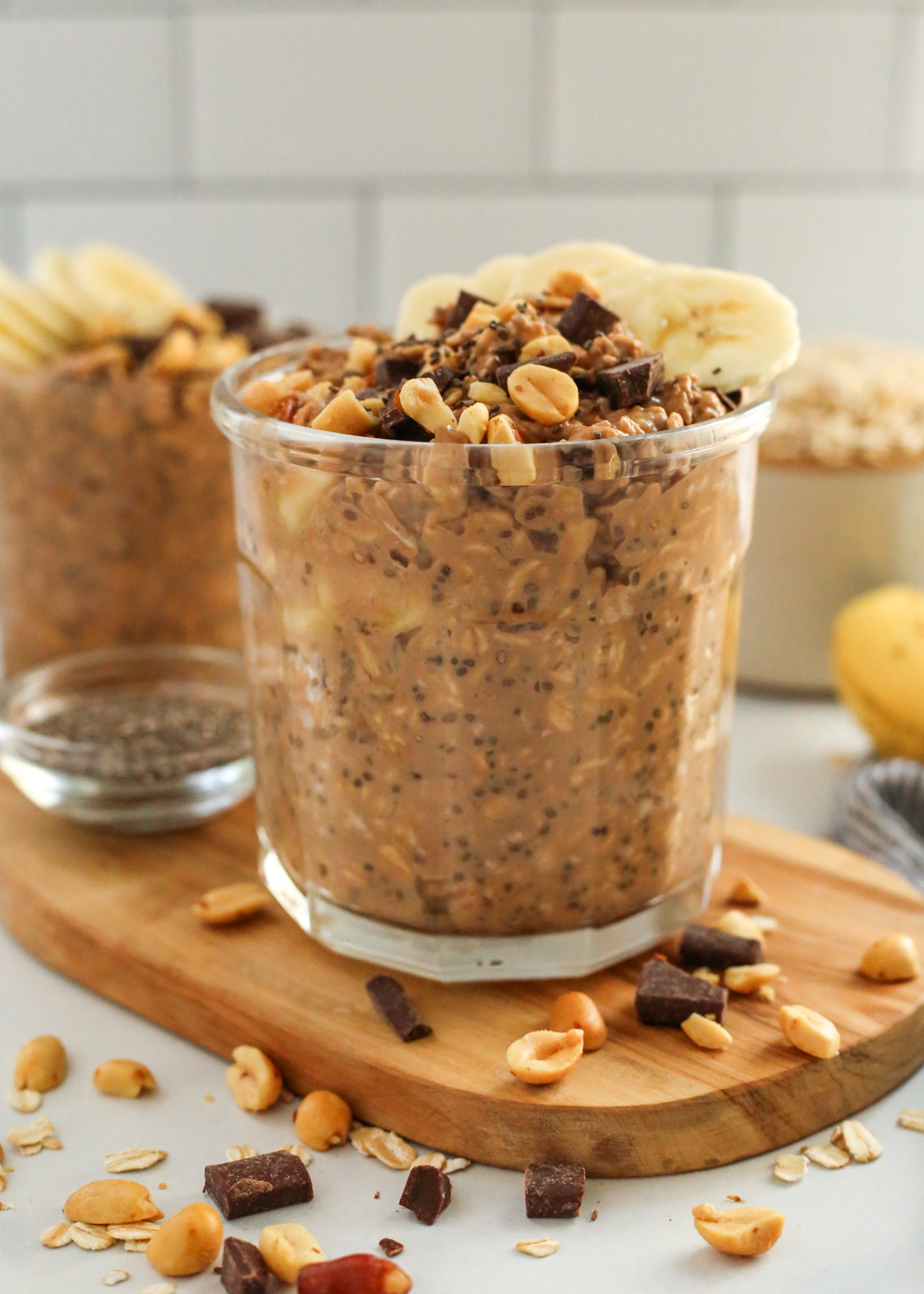 A glass mason jar holds a heaping portion of chocolate banana overnight oats, which are garnished with crushed peanuts, chocolate chunks, and slices of bananas, with a second jar visible in the background along with some of the included ingredients
