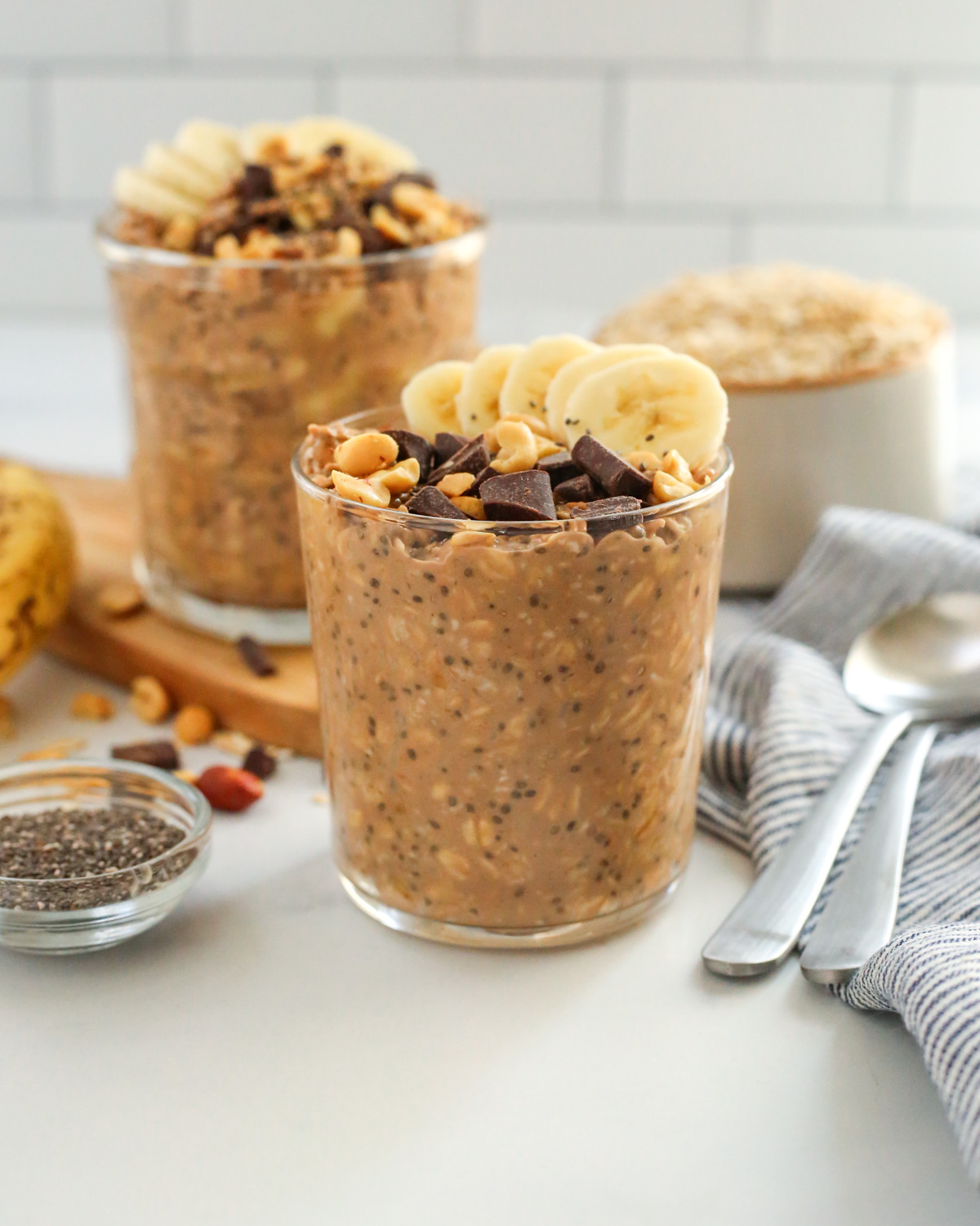 A glass cup containing chocolate banana overnight oats is displayed on a kitchen countertop, garnished with slices of banana, crushed peanuts and chocolate chunks, with a small ramekin of chia seeds and silver spoons resting on the side