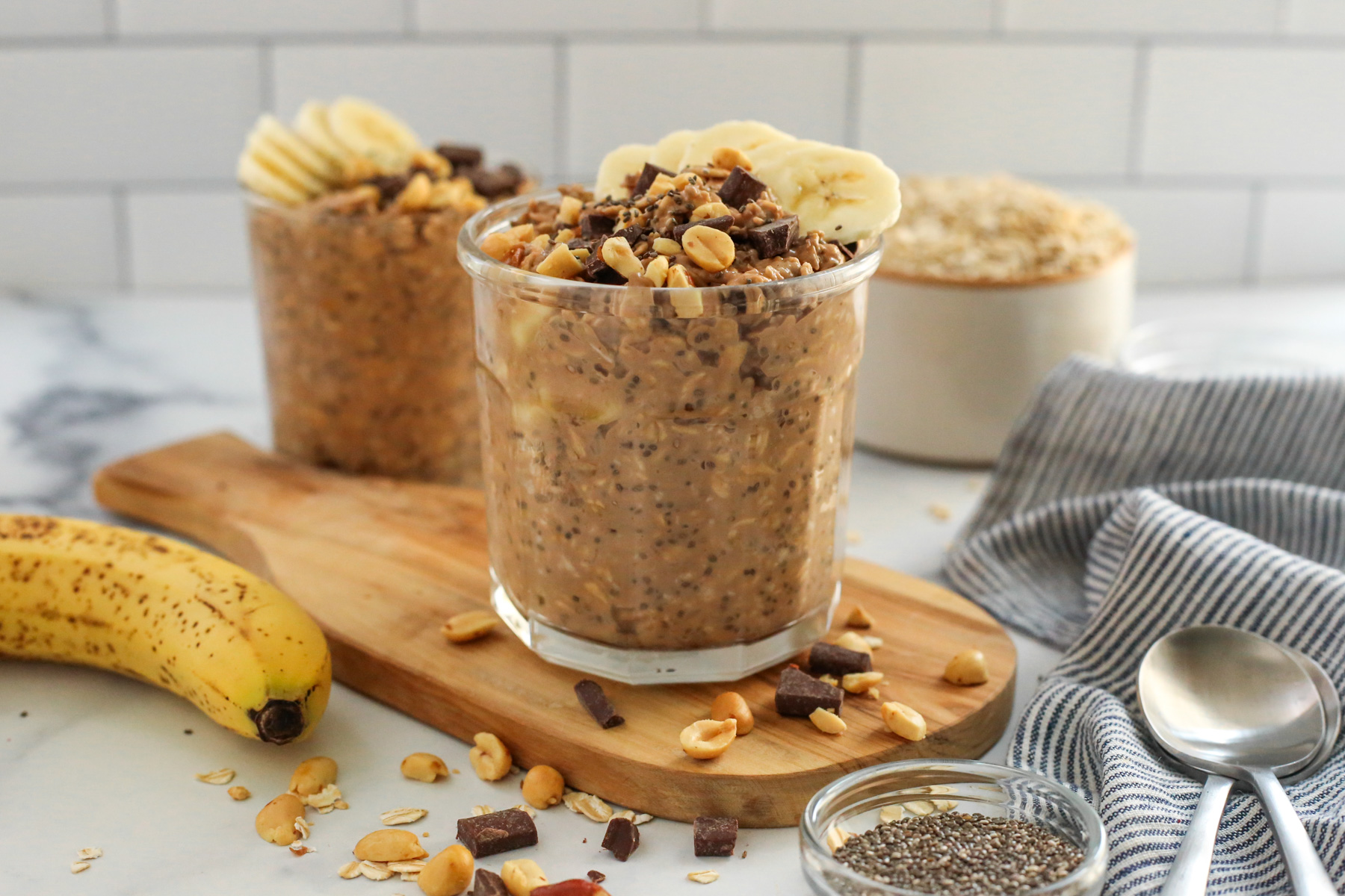 Kitchen countertop displaying a glass mason jar of chocolate banana overnight oats with a ripe banana, cup of uncooked oats, ramekin of chia seeds, and two silver spoons on a blue and white striped linen displayed nearby