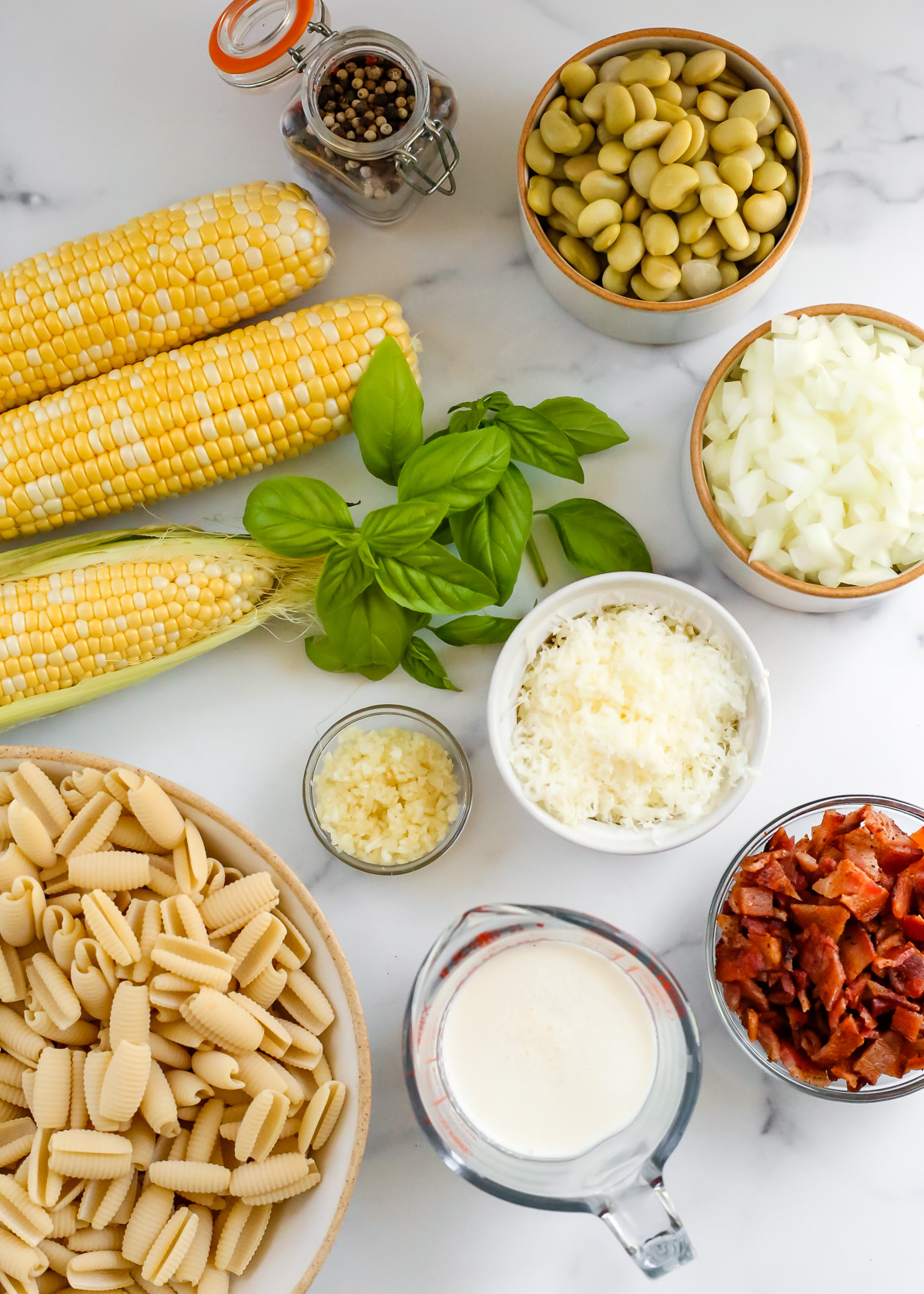 An assortment of ingredients on a white marble kitchen countertop, including ears of sweet corn, drained canned butter beans or lima beans, diced onions, sprigs of fresh basil leaves, black peppercorns, shredded mozzarella cheese, minced garlic, cooked bacon, a measuring cup with heavy cream, and uncooked pasta