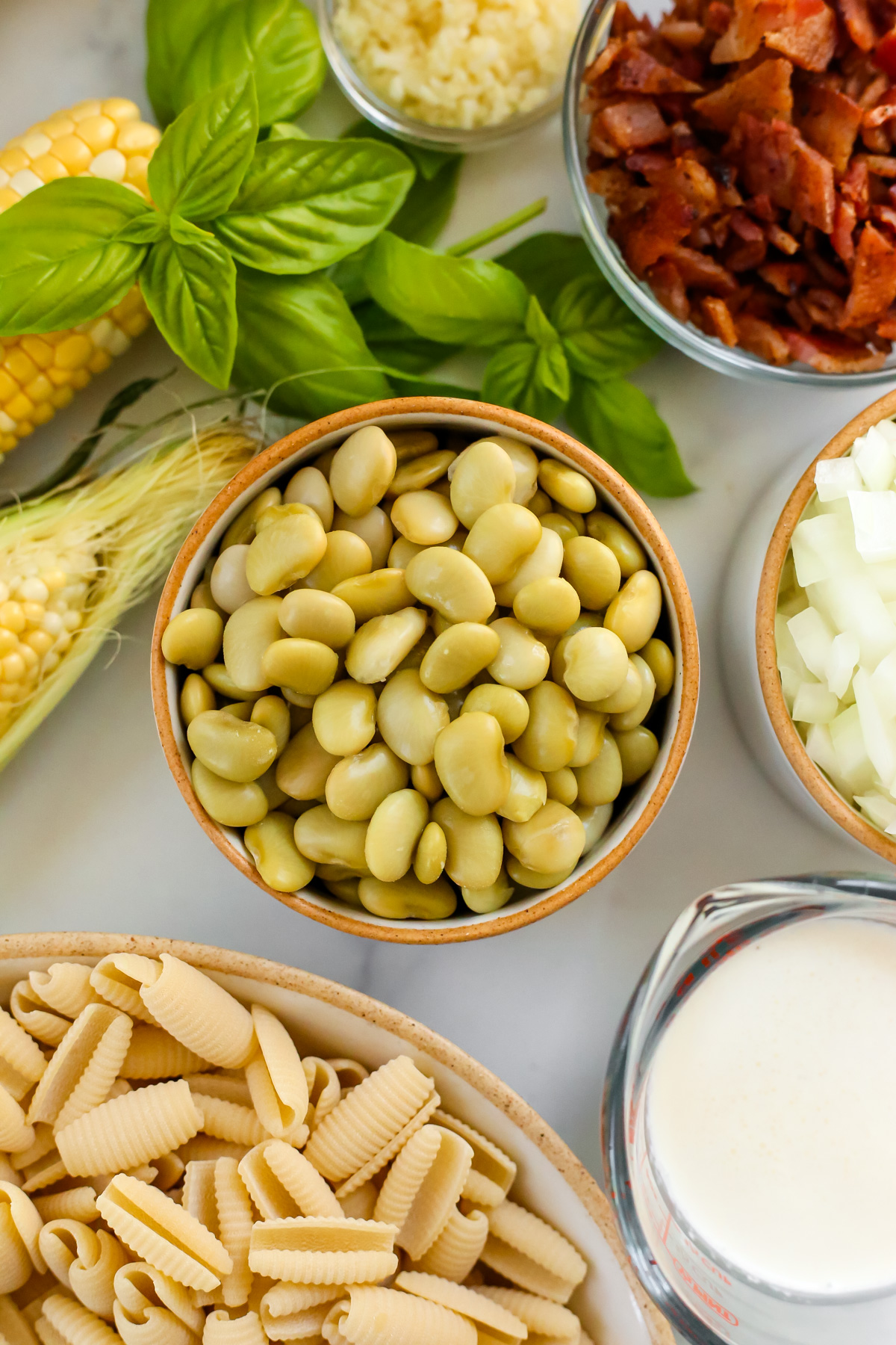 Overhead view of canned butter beans that have been drained and rinsed, shown in a small ceramic ramekin and surrounded by fresh basil, cooked bacon, diced onions, and an ear of fresh sweet corn