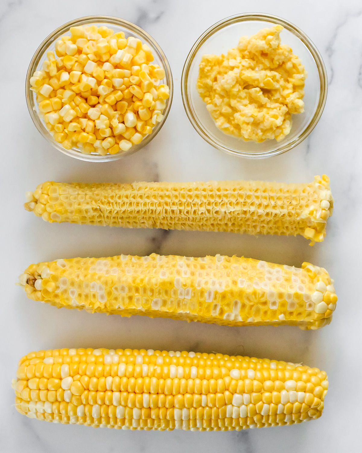 Overhead view of three corn cobs, showing the difference between a shucked ear of corn, a cob with the kernels sliced off, and a cob with the kernels milked. Two small glass ramekins show the corn kernels compared to the milked corn