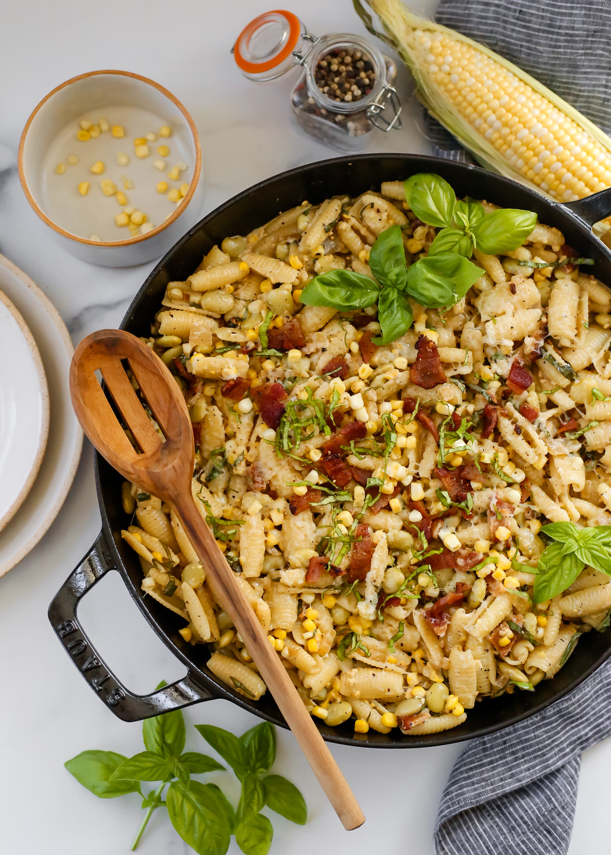A large Staub braising dish contains a summer pasta recipe with corn and a creamy sauce, plus bacon, sweet corn kernels, fresh basil, butter beans, and mozzarella cheese. The pan is garnished with sprigs of fresh basil leaves and a wooden serving spoon rests on the side