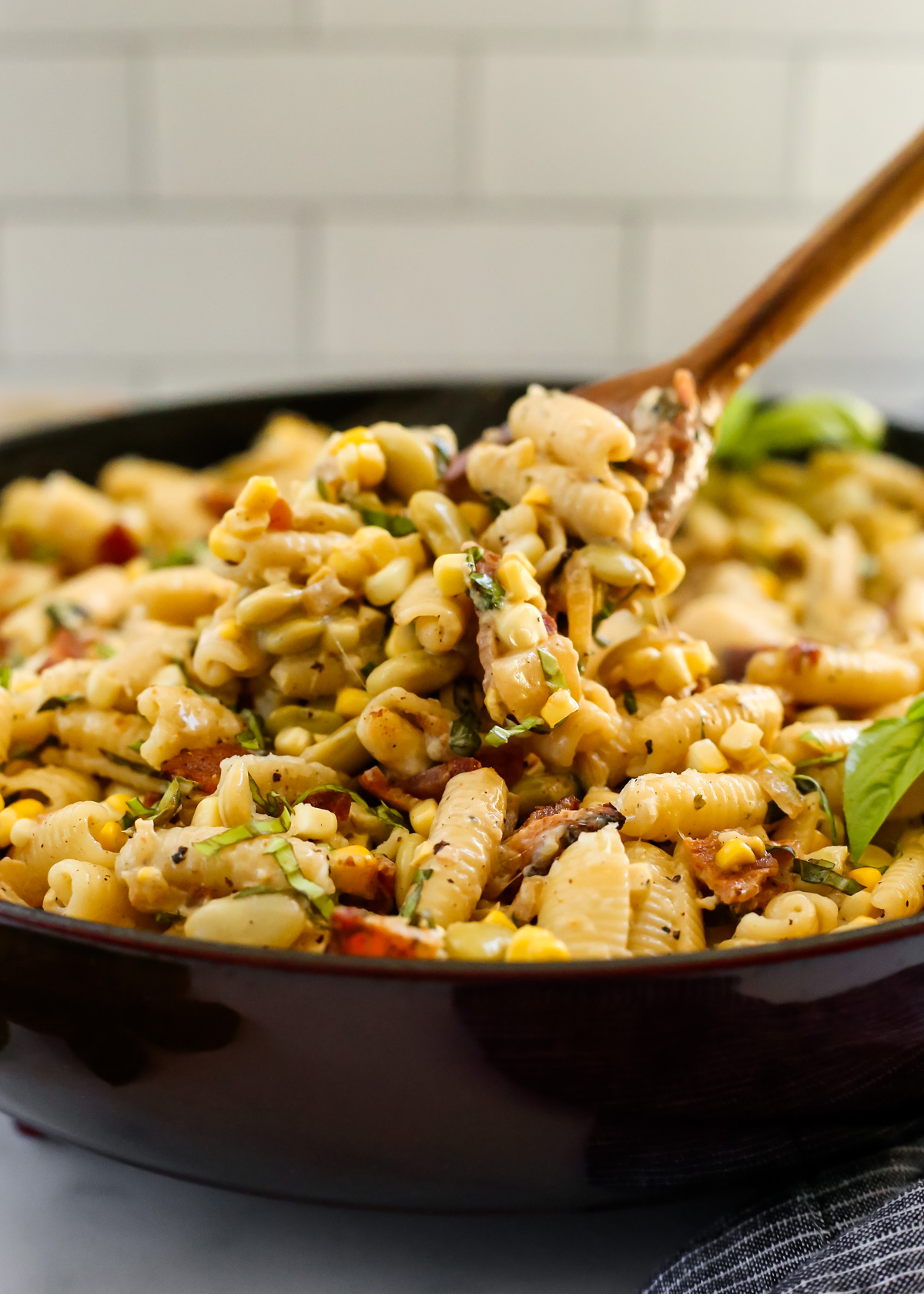 A wooden serving spoon lifts a heaping spoonful of a creamy corn pasta out of a braising dish filled to the brim with a saucy creamy pasta with visible chunks of bacon, fresh basil, and black peppers