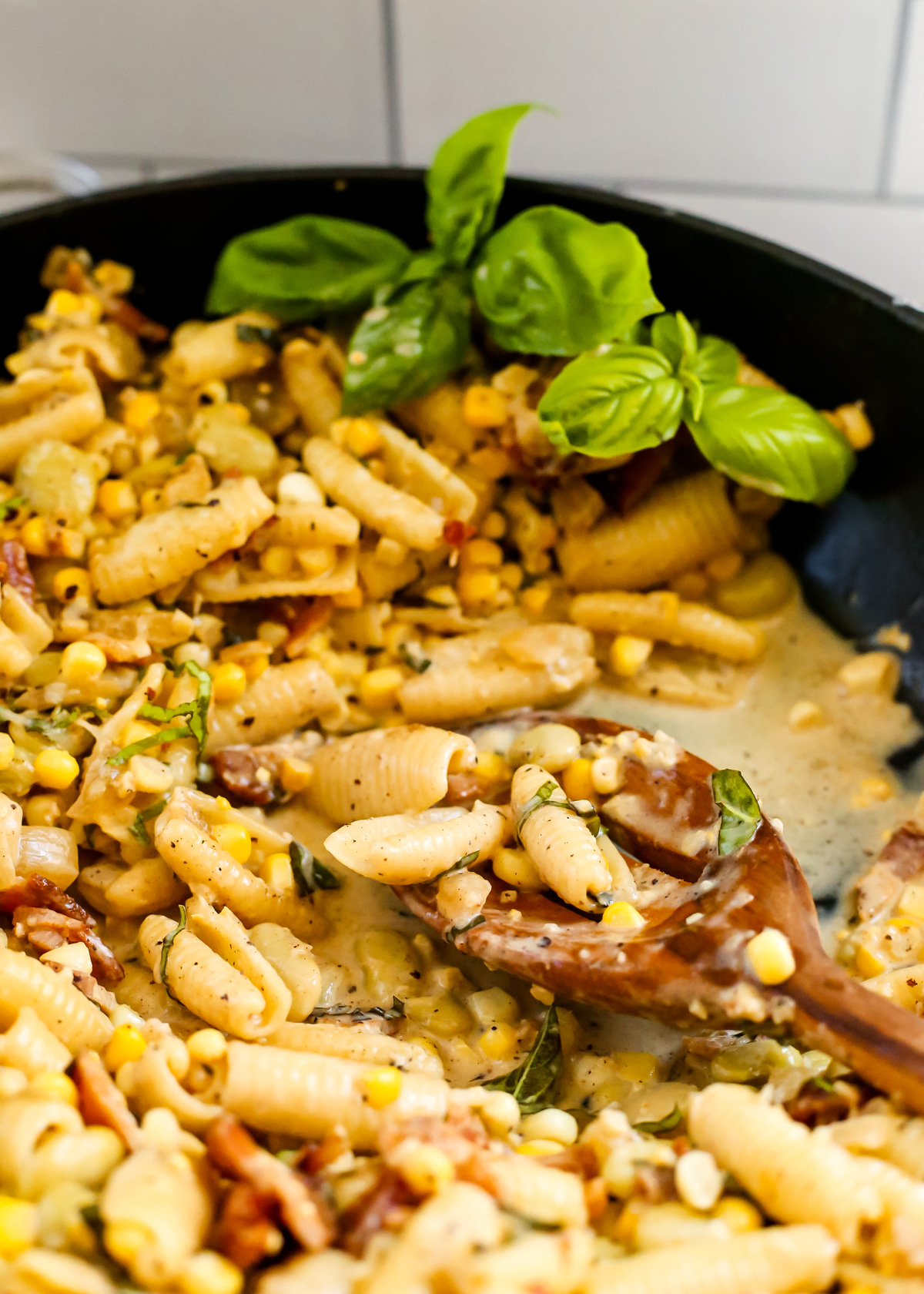 A view inside a large skillet used to make a creamy corn pasta recipe, with a wooden serving spoon stirring the contents that are coated in a creamy white sauce