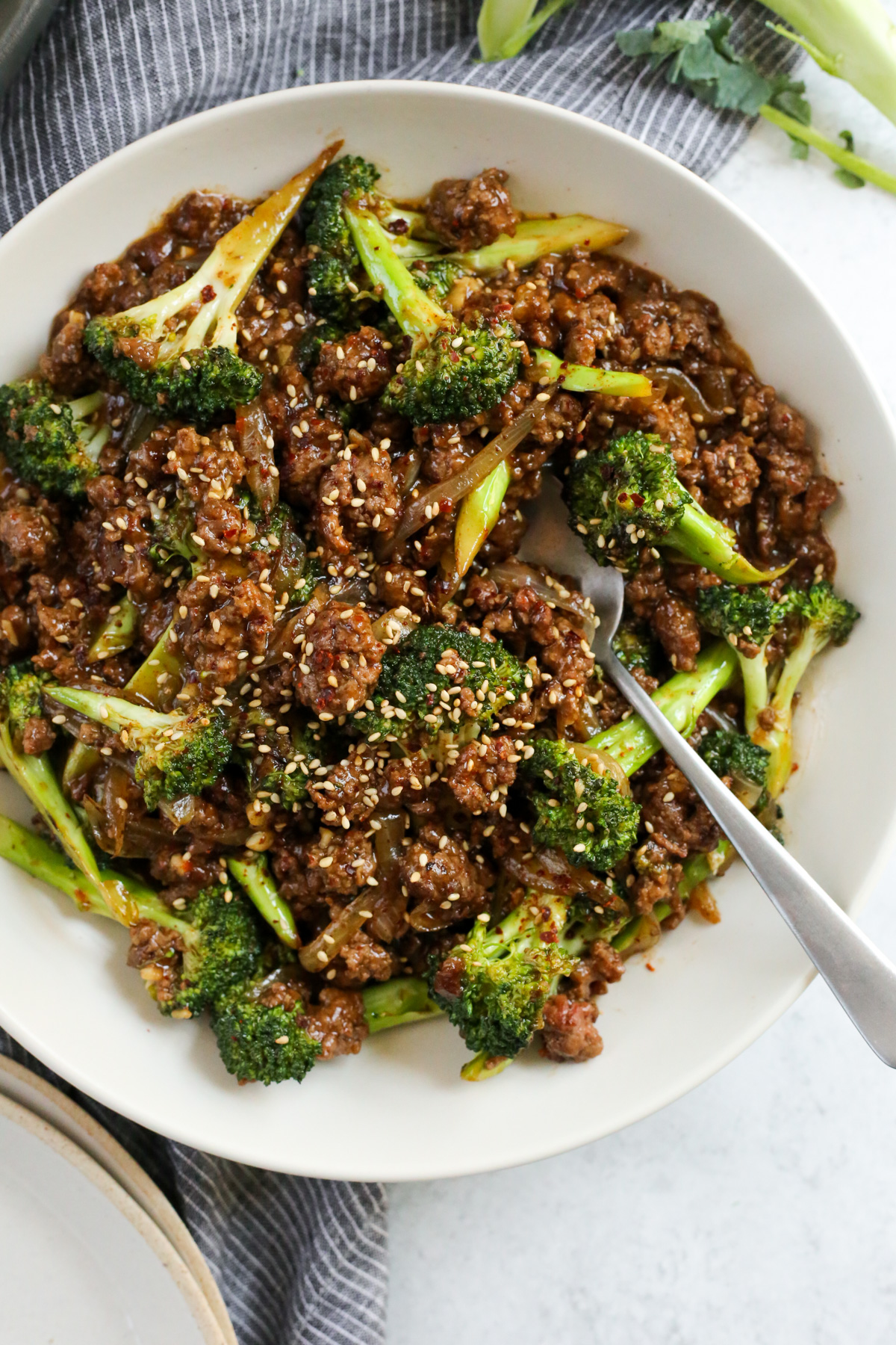 A silver serving spoon rests on the side of a bowl filled with a mixture of cooked ground beef, onions, and broccoli covered with a savory sauce and garnished with toasted sesame seeds