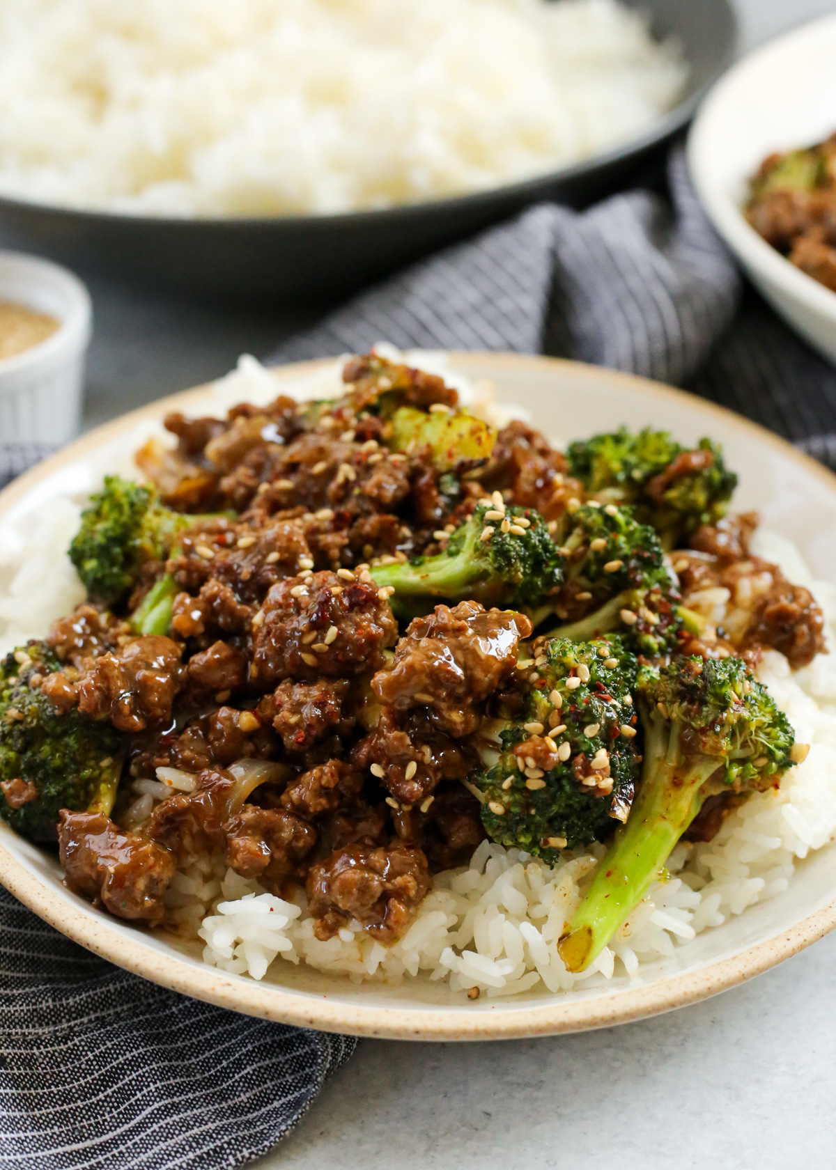 A side view of a plate filled with steamed white rice and covered with ground beef and broccoli in a silky savory sauce, garnished with toasted sesame seeds and ref pepper flakes