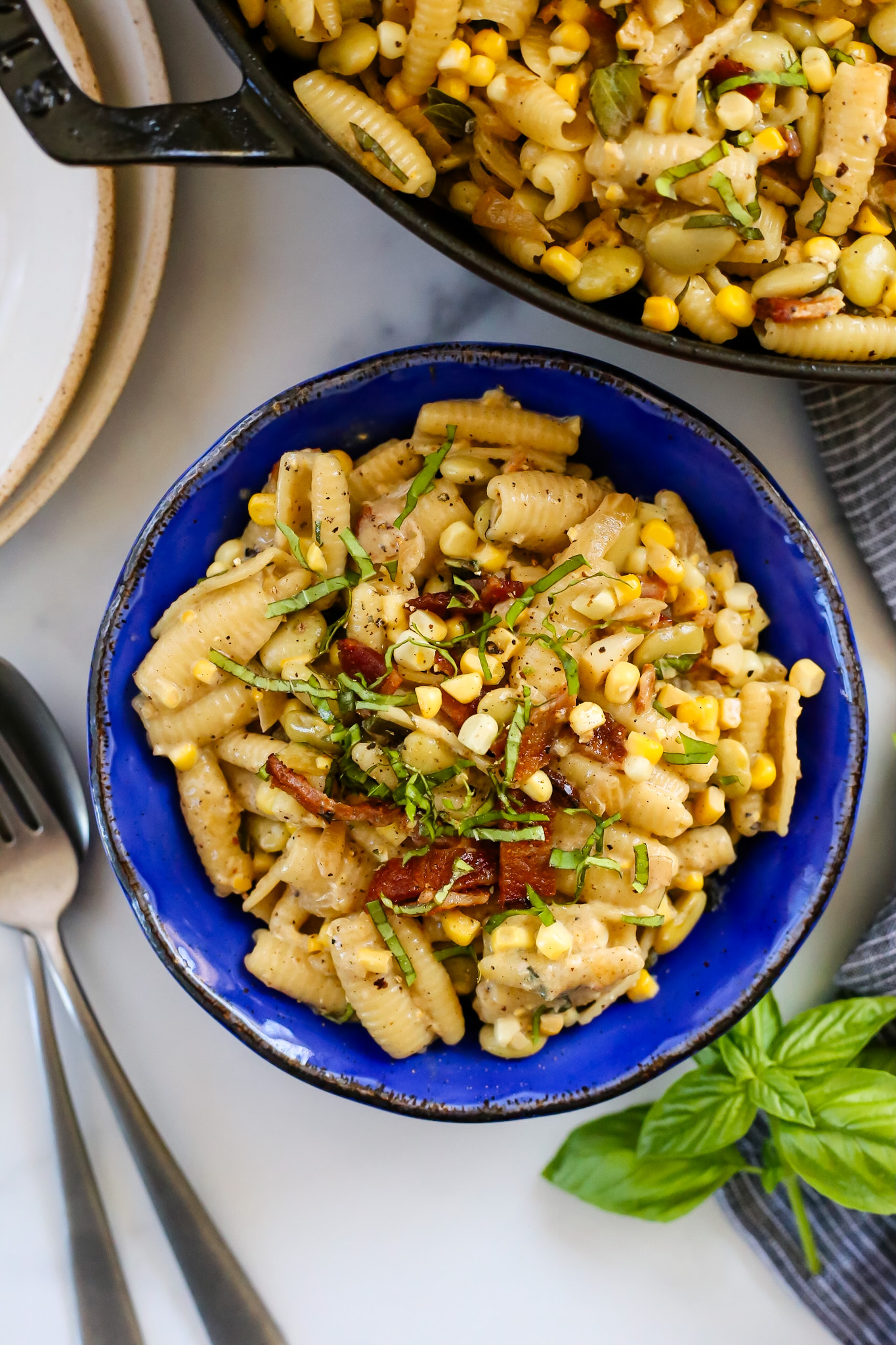 Overhead view of a creamy corn pasta recipe served in a blue glazed ceramic bowl, garnished with crumbles of bacon, fresh basil, and sweet corn kernels