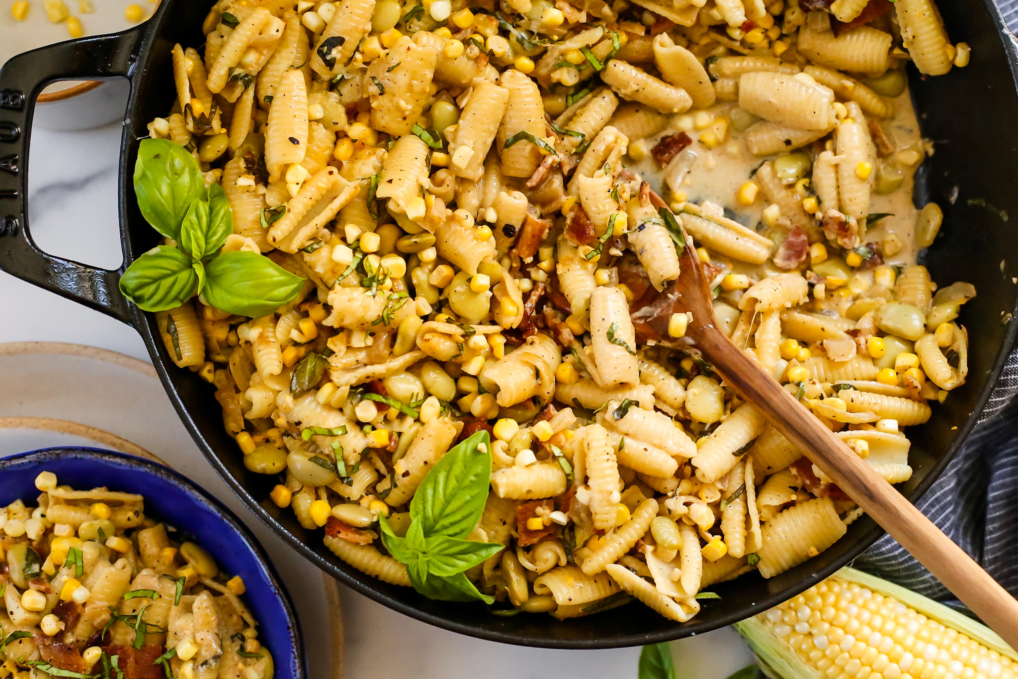 Overhead view of a large skillet containing a creamy corn pasta recipe with a small bowl served to the side, with both of them garnished with bacon crumbles, fresh basil, and cracked black pepper