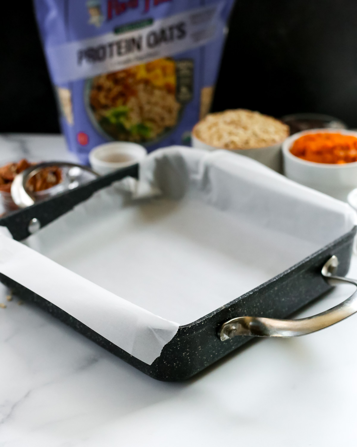 A square baking sheet with silver handles sits in front of the ingredients of making easy pumpkin oat bars, with a piece of white parchment paper lining the inside of the baking pan