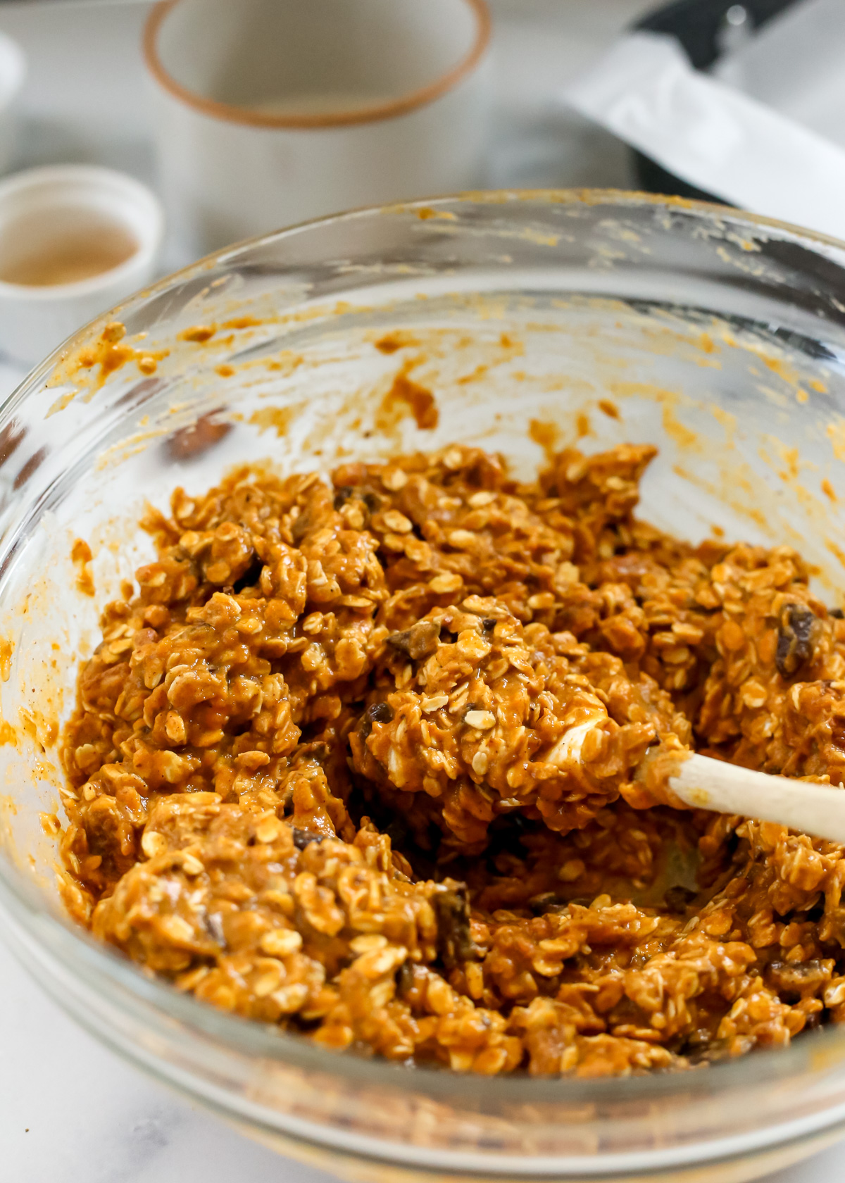 A small spatula rests on the side of a glass mixing bowls containing a mixture of oats, pureed pumpkin, brown sugar, pecans, and dark chocolate chunks, appearing bright orange in color and thick and sticky