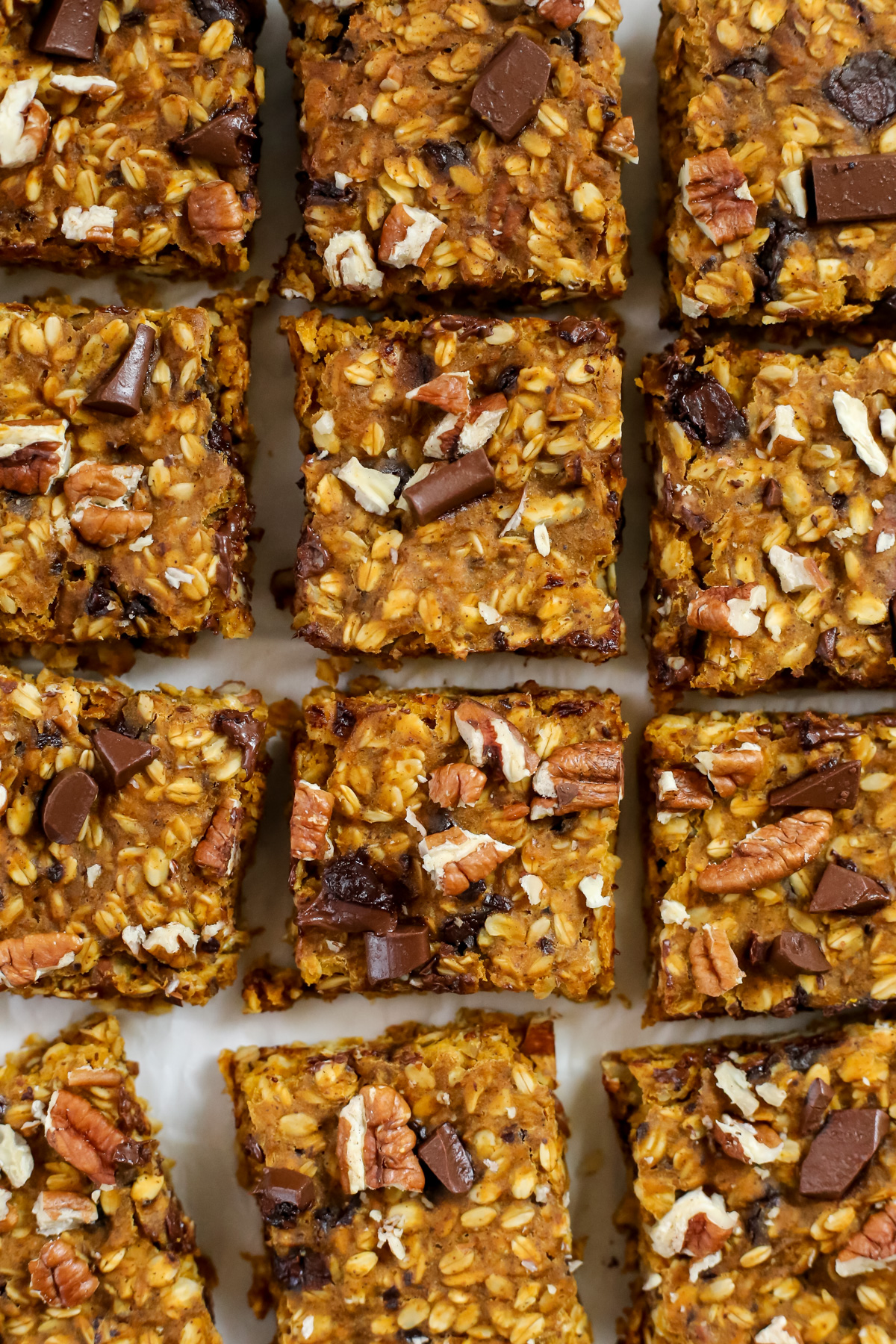 Overhead view of a batch of pumpkin bars topped with chunks of chocolate and pecan pieces, sliced and arranged on a piece of white parchment paper